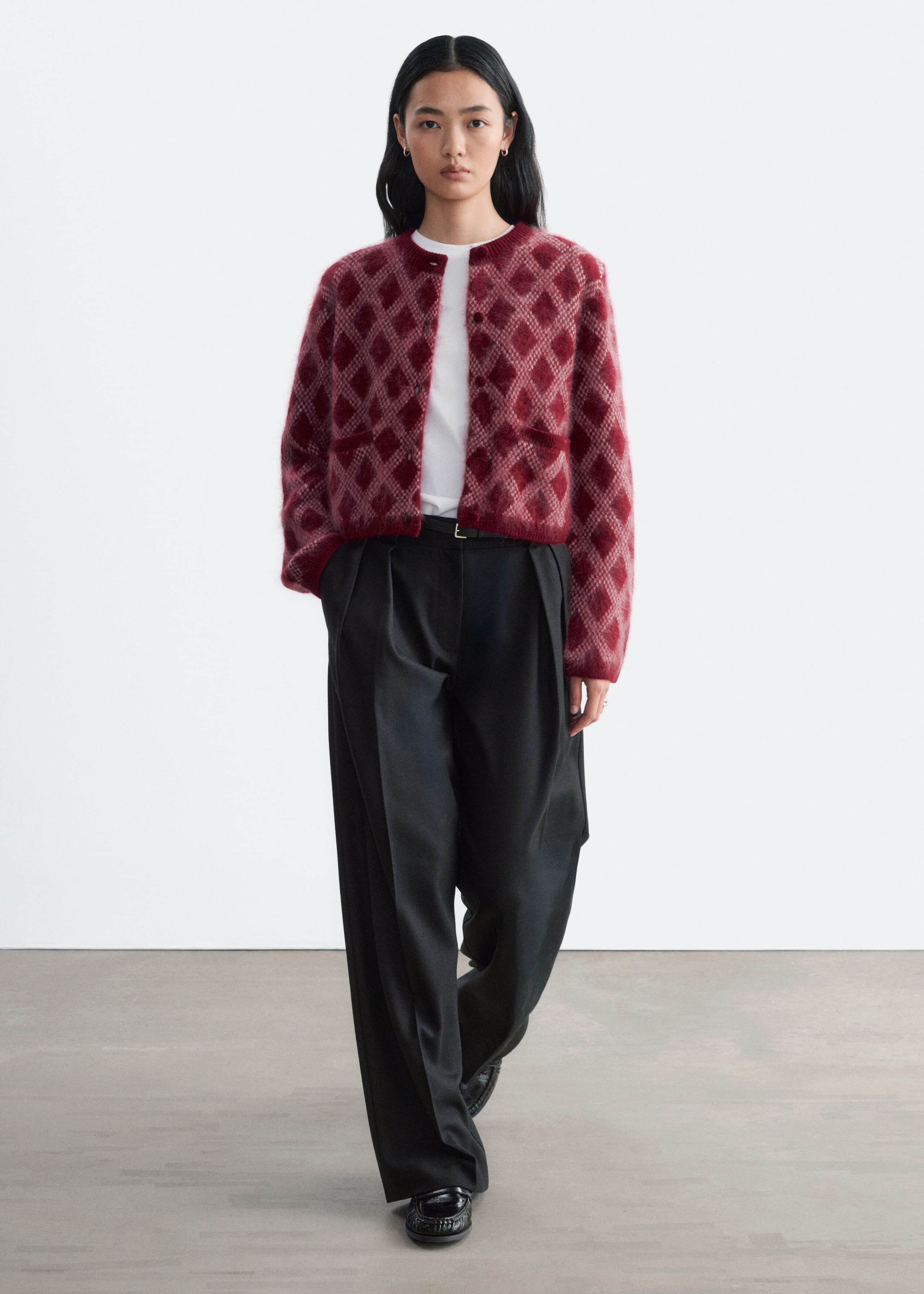 A fuzzy knit cardigan featuring a red and pink diamond pattern is worn open by the model over a white T-shirt and black wide-leg trousers. The cardigan features a structured shoulder silhouette, long sleeves, and a crew neckline.