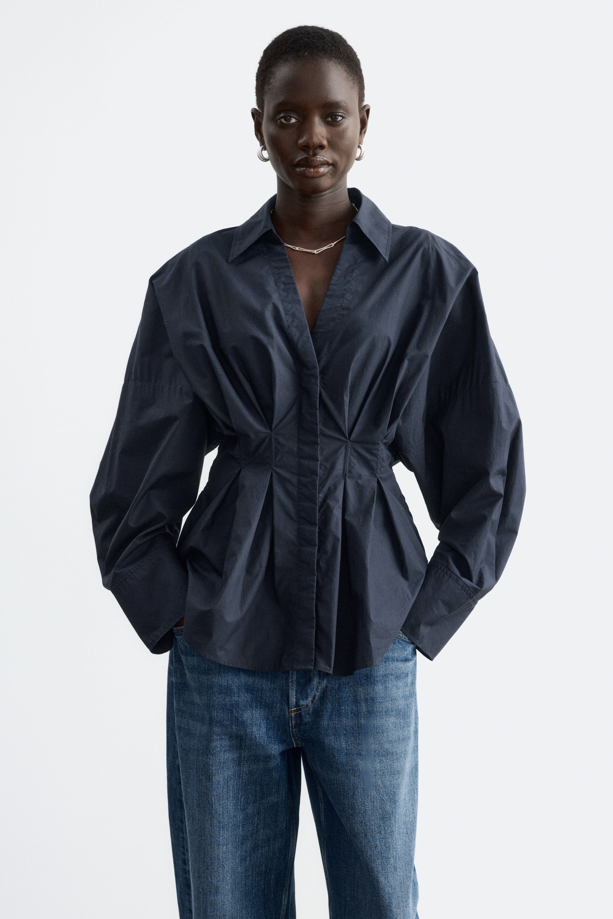 Visualizza l'immagine ingrandita: La modella indossa una camicia in cotone blu navy scuro, caratterizzata da una vita aderente con dettagli a pieghe, maniche voluminose e collo aperto. La camicia è abbinata a jeans blu, mentre la modella tiene le mani in tasca.