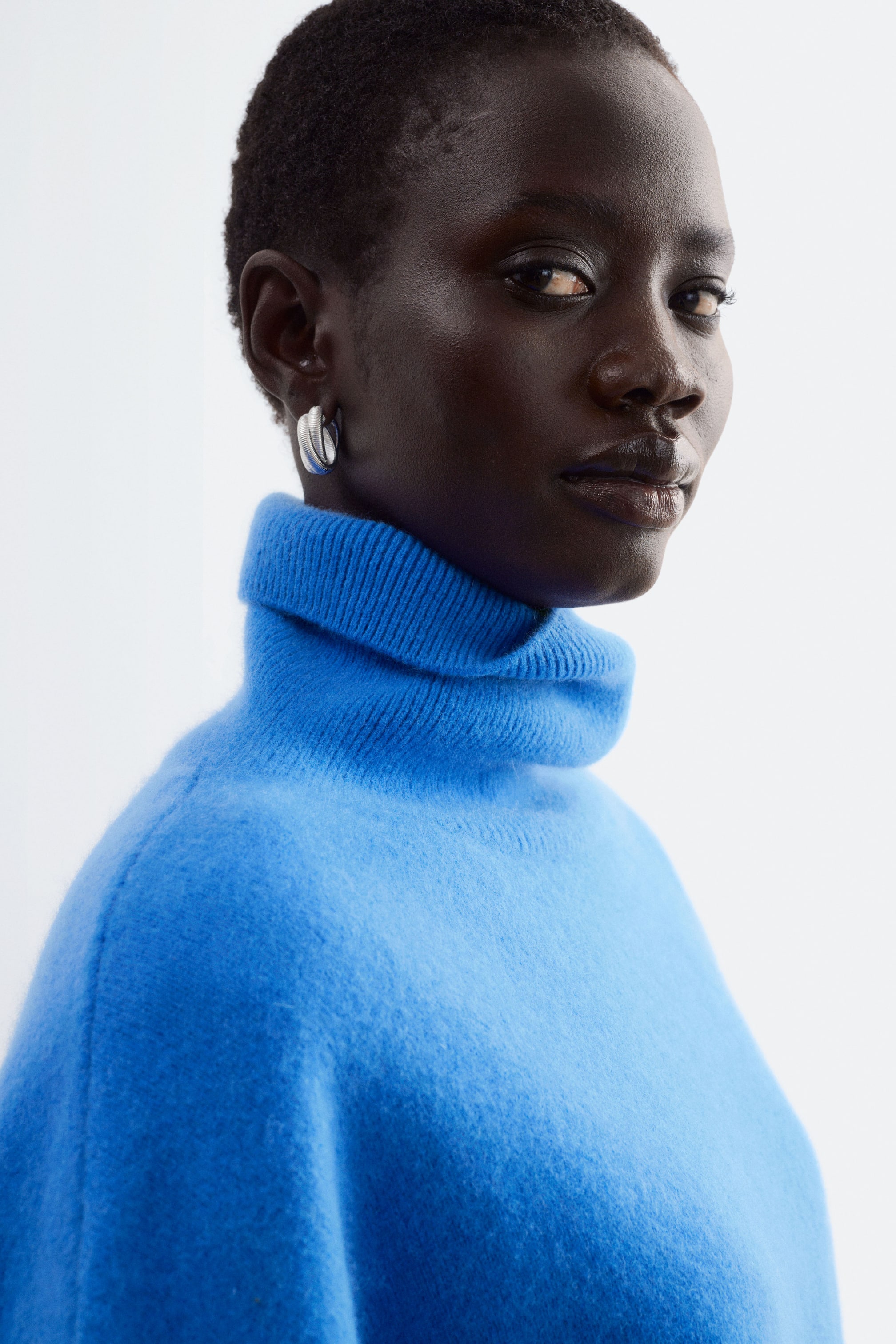 Agrandir l'image: Un pull en laine mérinos bleu vif avec un col roulé épais à revers côtelé est porté par le modèle, assorti à une boucle d’oreille créole torsadée ton argenté avec une surface texturée.