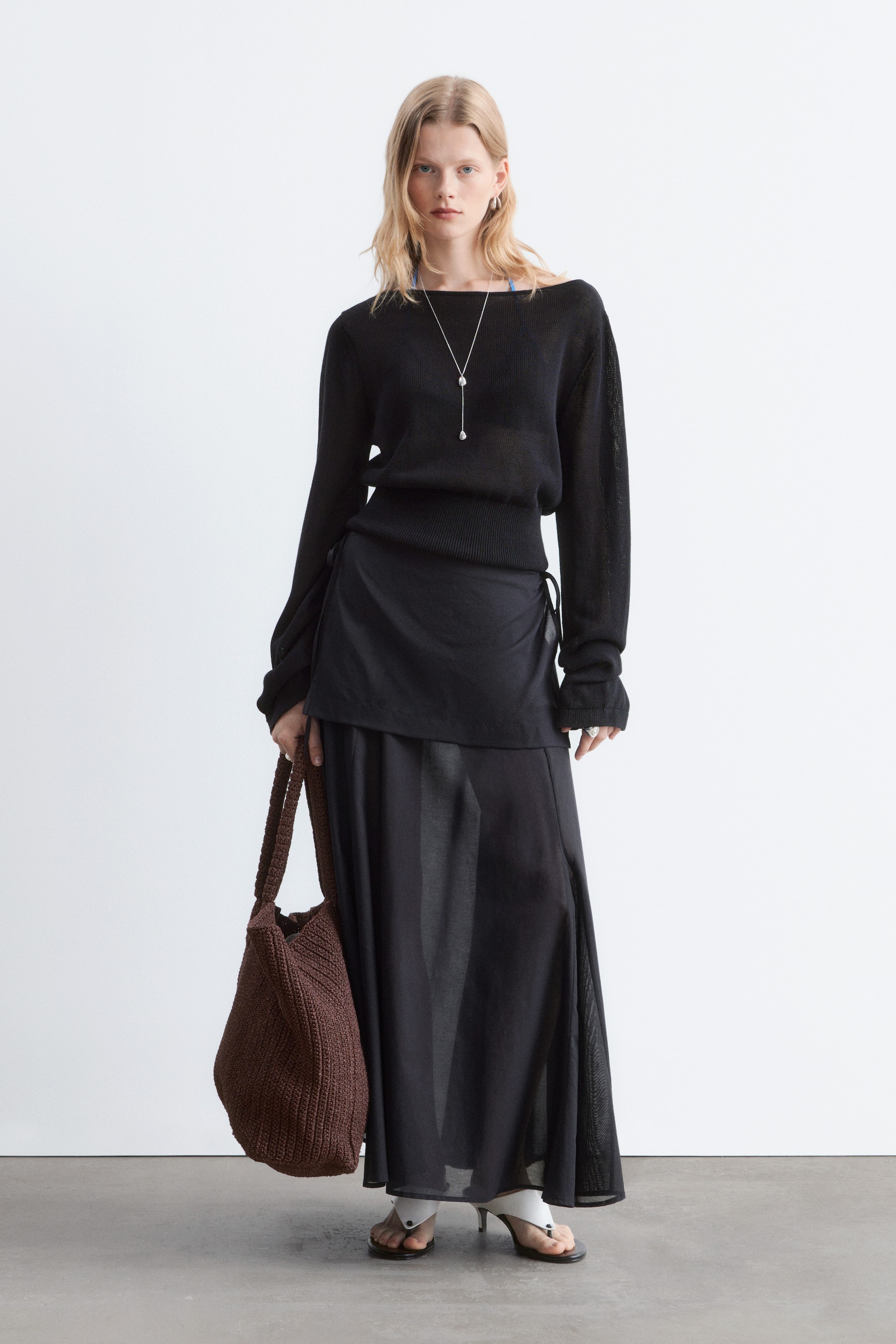 Grotere afbeelding bekijken: Een zwarte, doorschijnende top met lange mouwen en een boothals, voorzien van geribbelde manchetten en zoom, wordt frontaal gepresenteerd. Deze is gestyled met een zwarte, doorschijnende maxi-rok met een split aan de zijkant en een gelaagd zwart wikkelpaneel. Een zilveren Y-ketting met druppelvormige hangers siert de halslijn, een bruine, gevlochten hobo-tas wordt gedragen, en witte teenslippers worden gedragen.