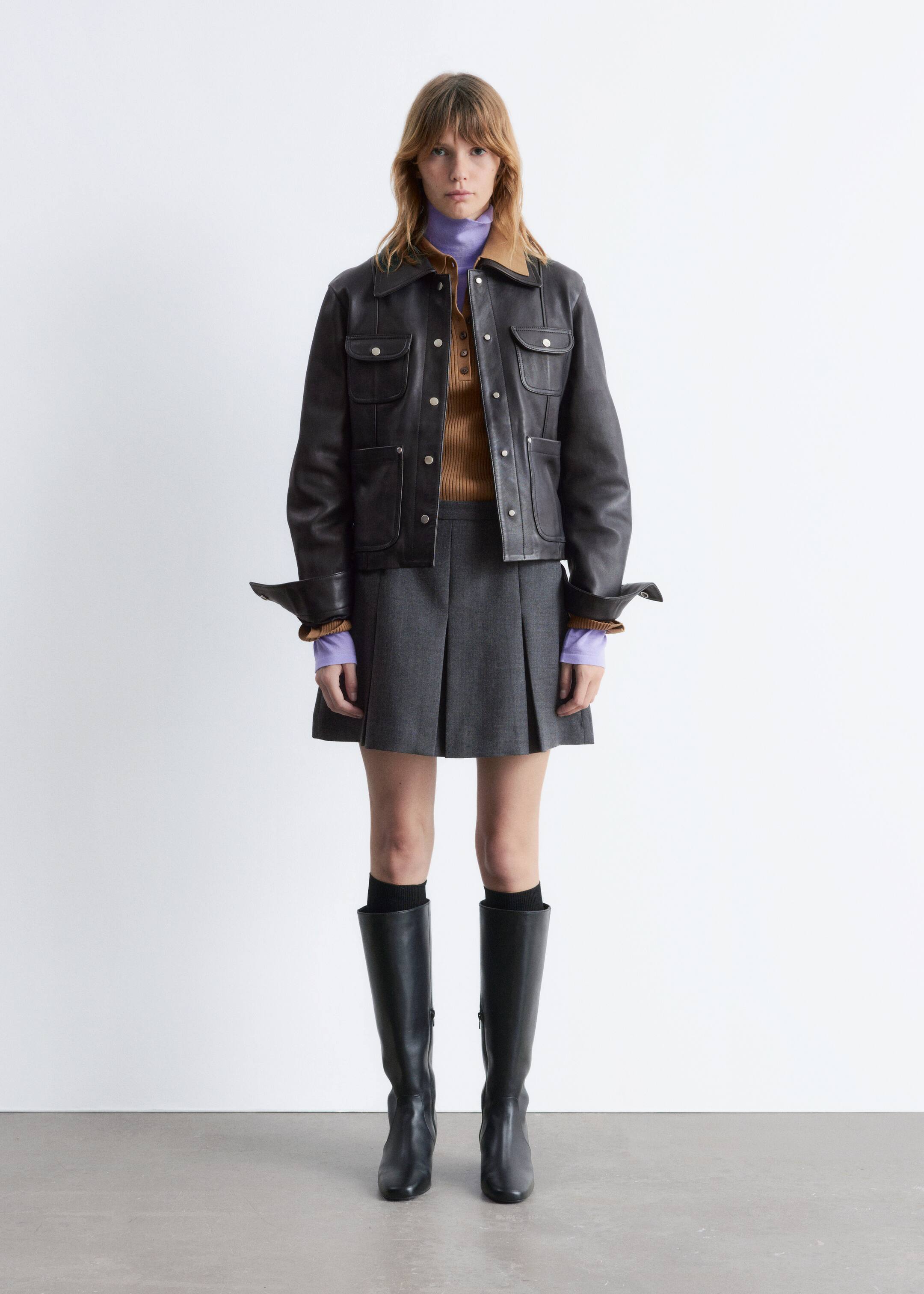 Se større billede: Et forfra-vendt outfit bliver præsenteret, og det har en mørkebrun jakke med en beige underkrave, fire klaplommer med trykknaplukning og en front snap slåkant. Jakkenes manchetter er slået op, hvilket afslører de ribbede manchetter på en brun henley-top med en lilla turtleneck, som bæres under en grå plisseret mini nederdel, sorte knæhøje støvler og sorte sokker.
