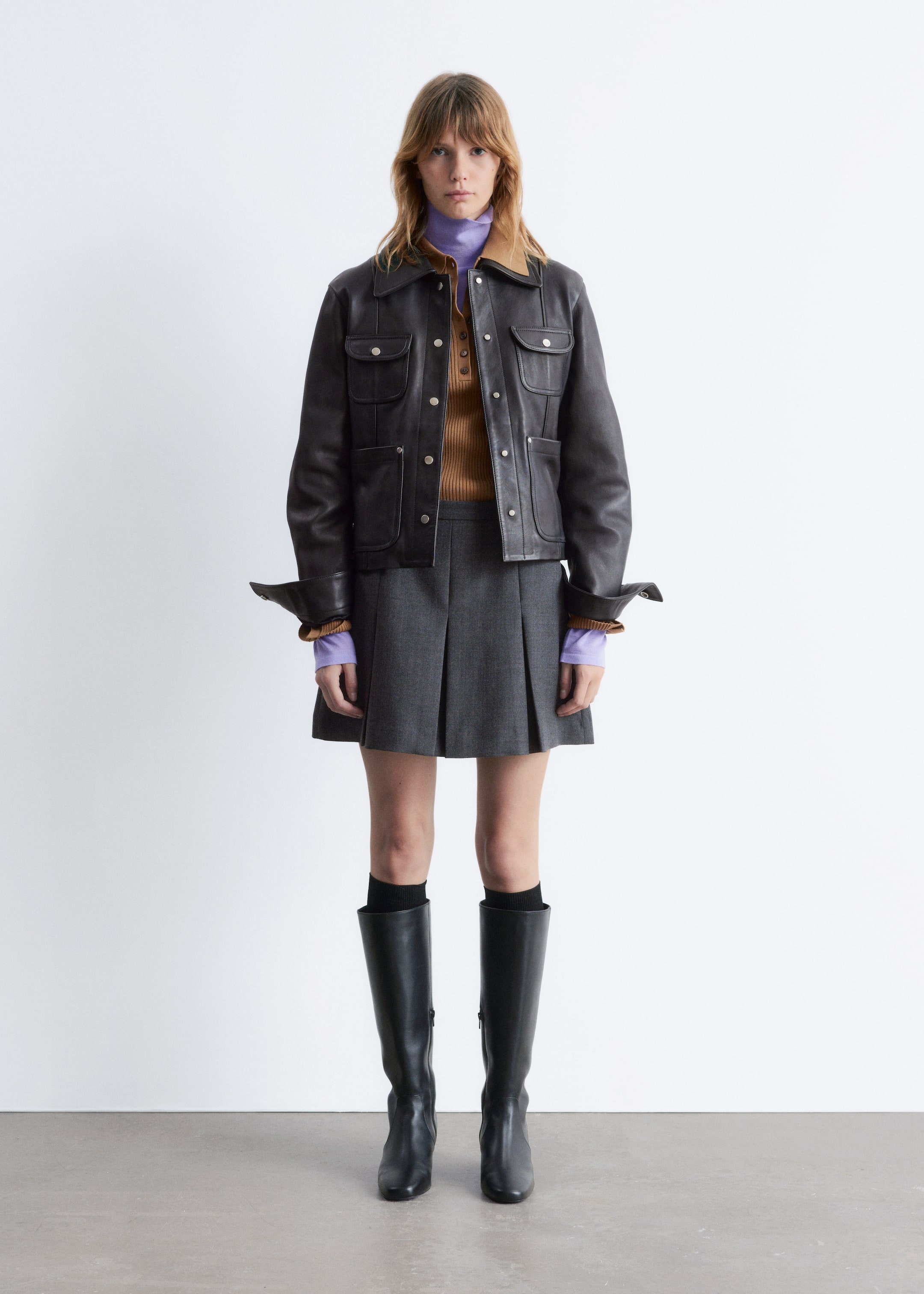 Visa större bild: En framåtvänd outfit presenteras, och den har en mörkbrun jacka med en beige underkrage, fyra ficklock med knapplås och en knäppning framtill. Jackans manschetter är uppvikta och avslöjar de ribbade manschetterna på en brun henley-topp med en lila polokrage, som bärs under med en grå plisserad minikjol, svarta knähöga stövlar och svarta strumpor.