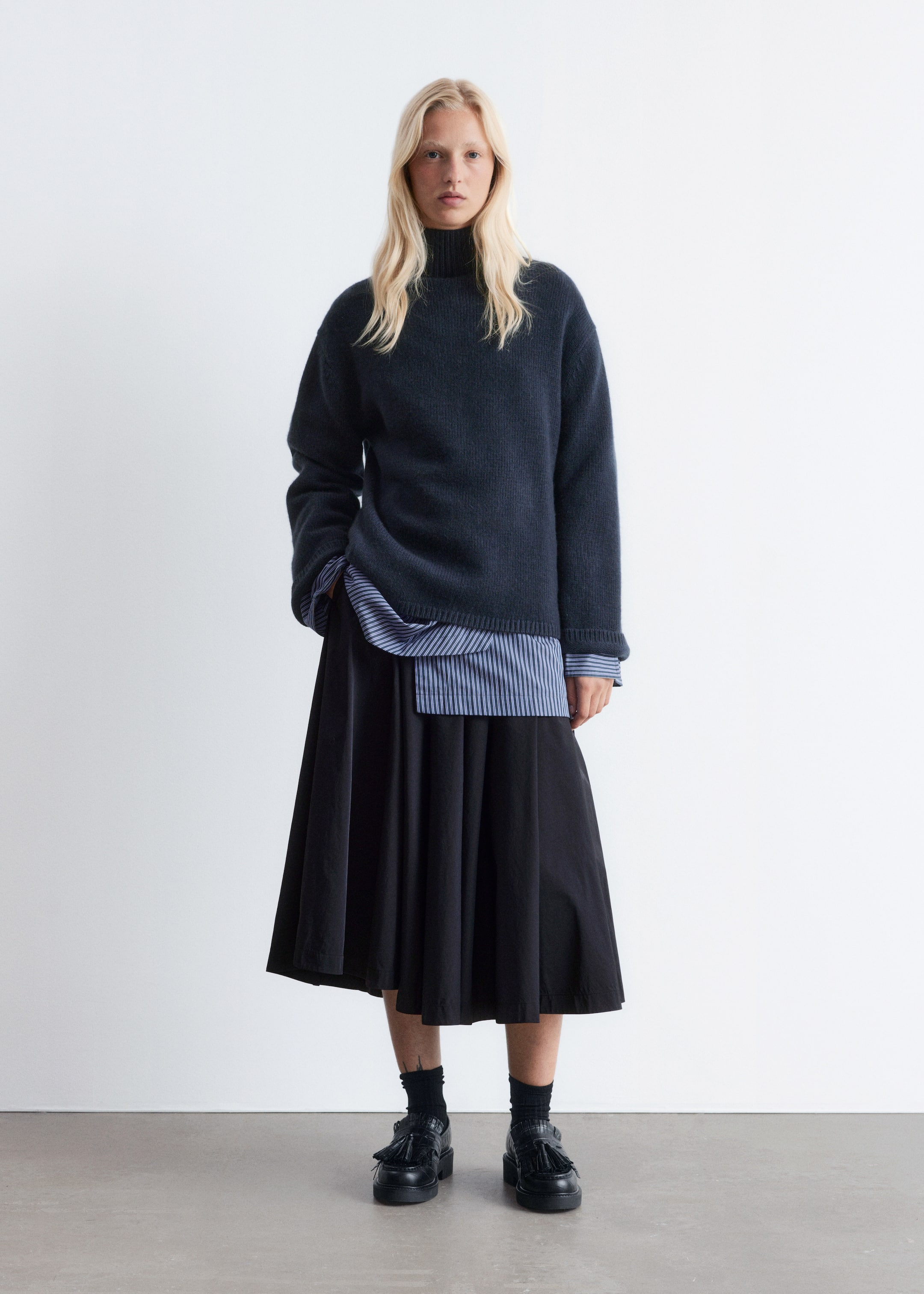 Grösseres Bild ansehen: Das Produkt ist ein dunkelblauer, locker gestrickter Rollkragenpullover, der über einem blau-weiß gestreiften Hemd getragen wird, dessen Saum und Manschetten sichtbar darunter hervorragen. Ein schwarzer, knielanger Midirock ergänzt das Oberteil und reicht bis zur Mitte der Waden, während schwarze Loafer mit Quasten und dicken Sohlen das Ensemble vervollständigen, wobei schwarze Socken darüber hervorschauen.