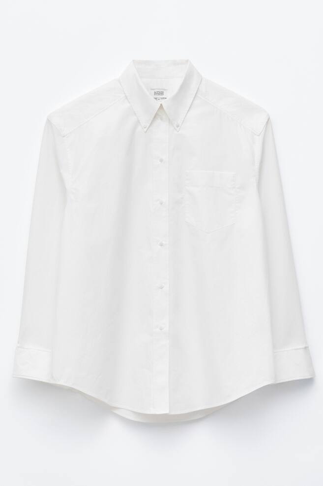 A white, long-sleeved button-down shirt is shown from the front, featuring a classic button-down collar, a central button placket, and a single chest patch pocket. The shirt has a relaxed fit, with wide cuffs on the sleeves and a curved hemline. - 1
