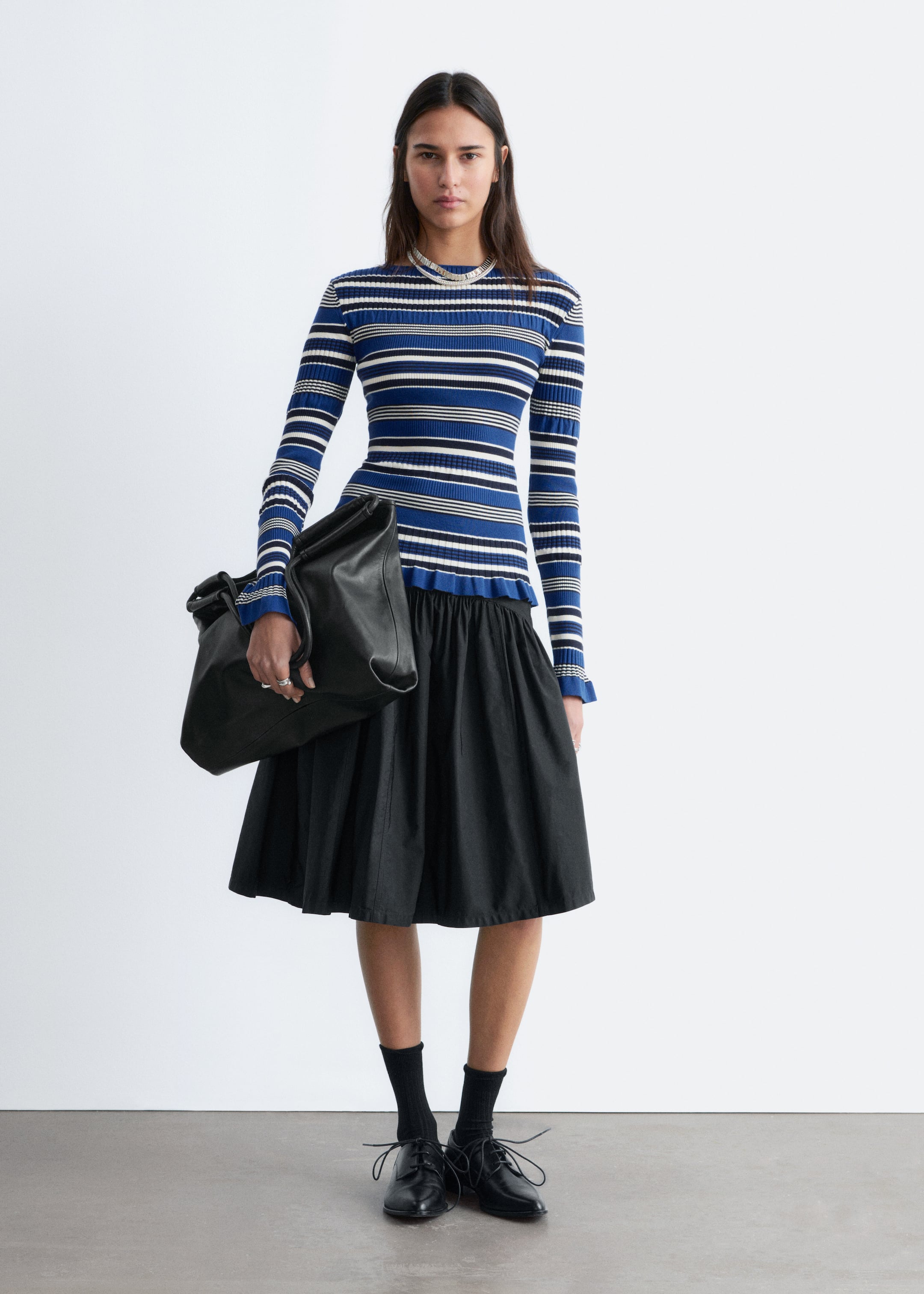 Zobrazit větší obrázek: Přiléhavý, dlouhý rukáv, žebrovaný pletený top s modrými, bílými a černými vodorovnými pruhy, s volánovými manžetami a volánovým lemem. Je stylizován s černou nabíranou midi sukní, černou volnou koženou taškou přes rameno a černými šněrovacími derby botami, které jsou nošeny s žebrovanými ponožkami.