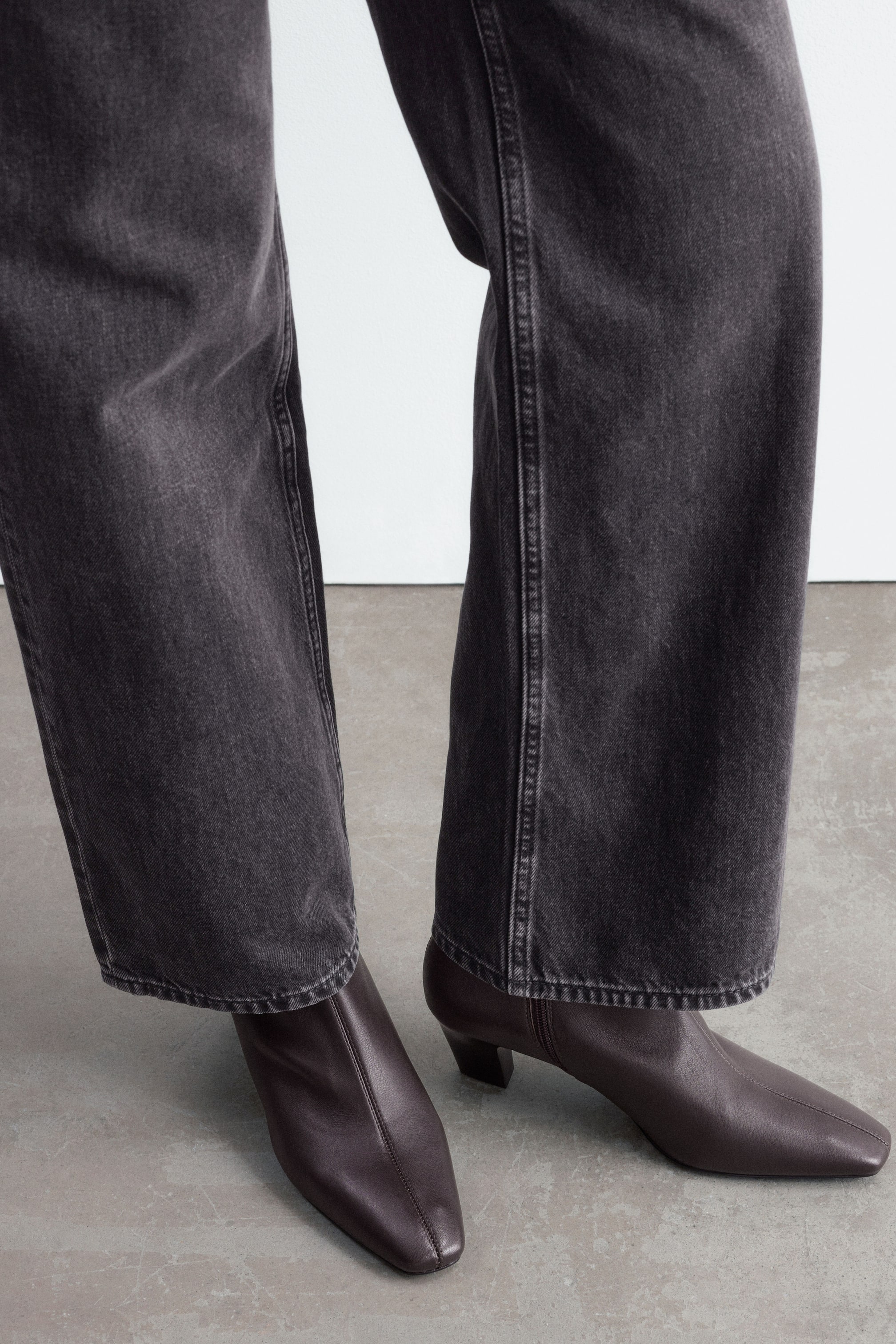 Ver imagen más grande: La modelo lleva unas botas tobilleras de cuero marrón oscuro con puntera cuadrada, costura central delantera y tacón bajo de bloque curvado. Las botas se combinan con unos vaqueros anchos de color gris oscuro.