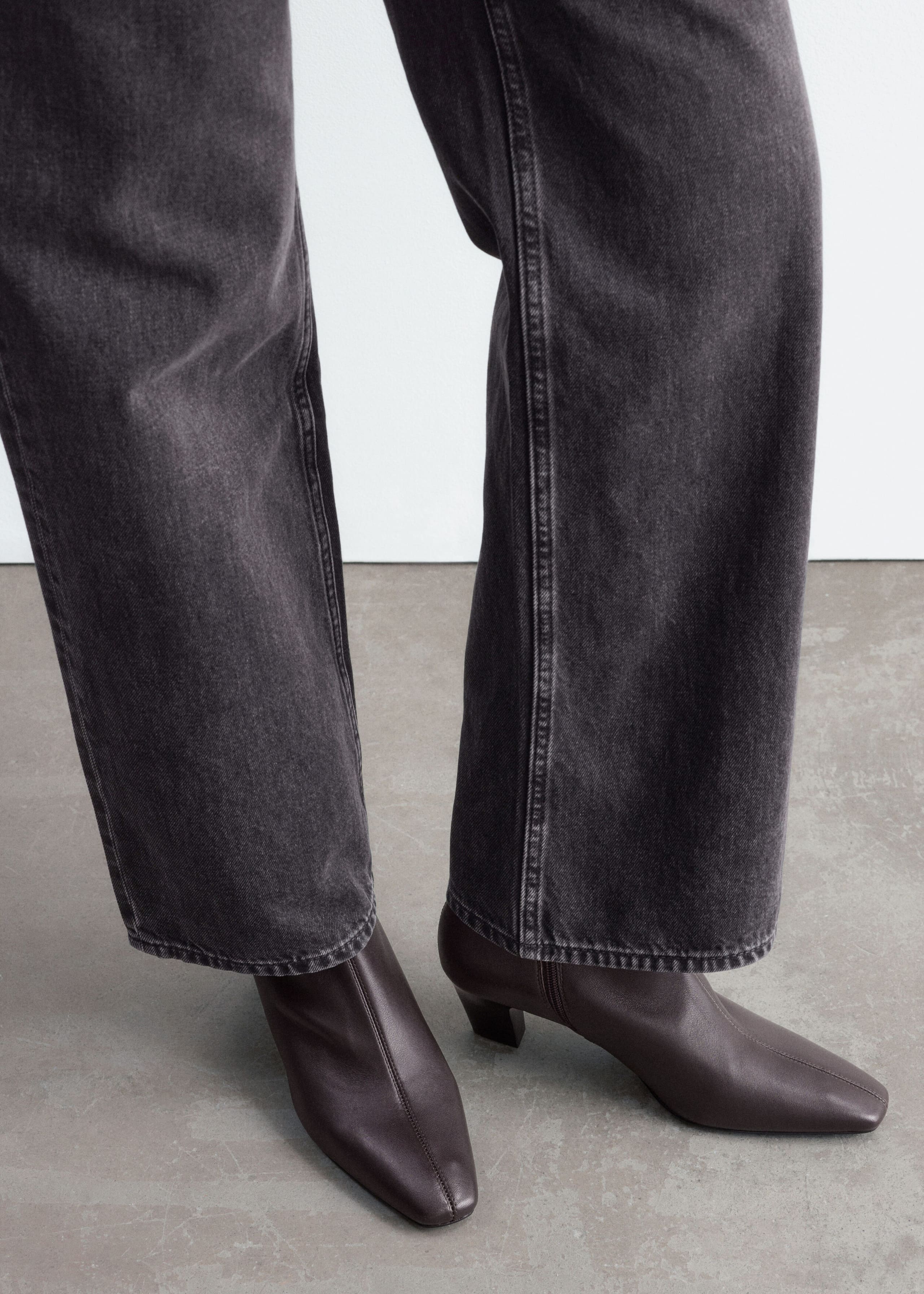Gli stivaletti in pelle marrone scuro, caratterizzati da punta squadrata, cucitura centrale sul davanti e tacco basso a blocco dalla forma curva, sono indossati dalla modella. Gli stivaletti sono abbinati a jeans denim grigio scuro a gamba larga.