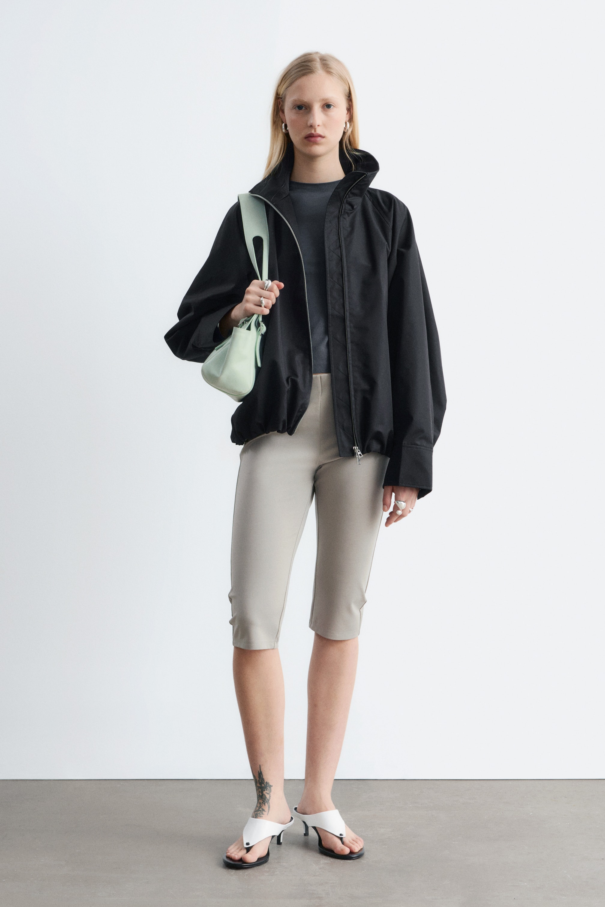 Ingrandisci l'immagine: La giacca nera presenta un colletto alto, maniche voluminose raccolte ai polsini e un orlo elasticizzato. Una zip frontale color argento corre al centro, con la giacca che si sovrappone a un top grigio scuro.