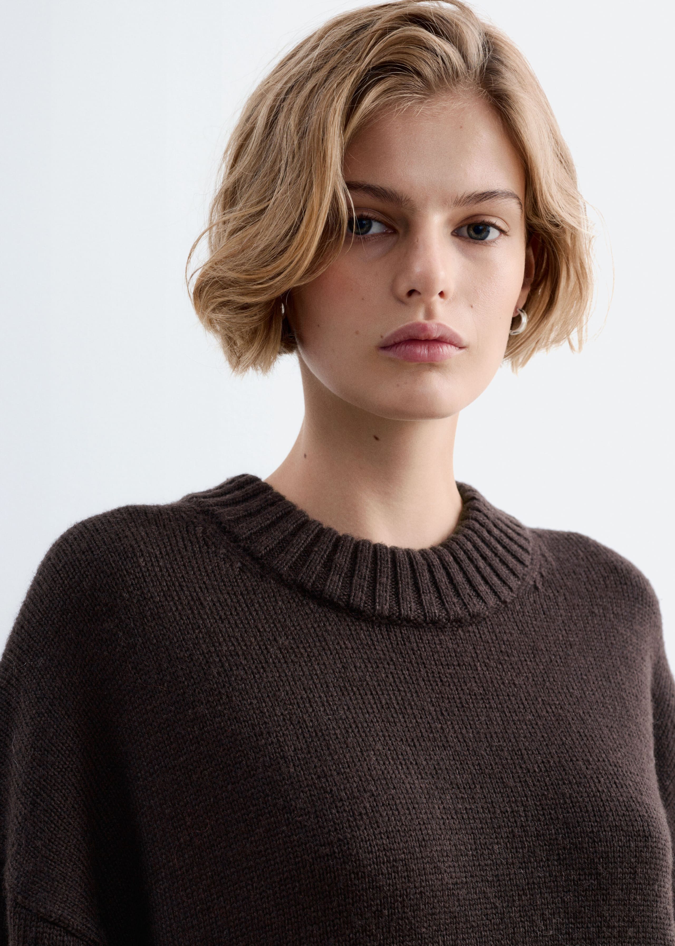 An oversized wool sweater in dark brown is being worn by the model, featuring a thick ribbed crew neckline and a fine-knit texture.