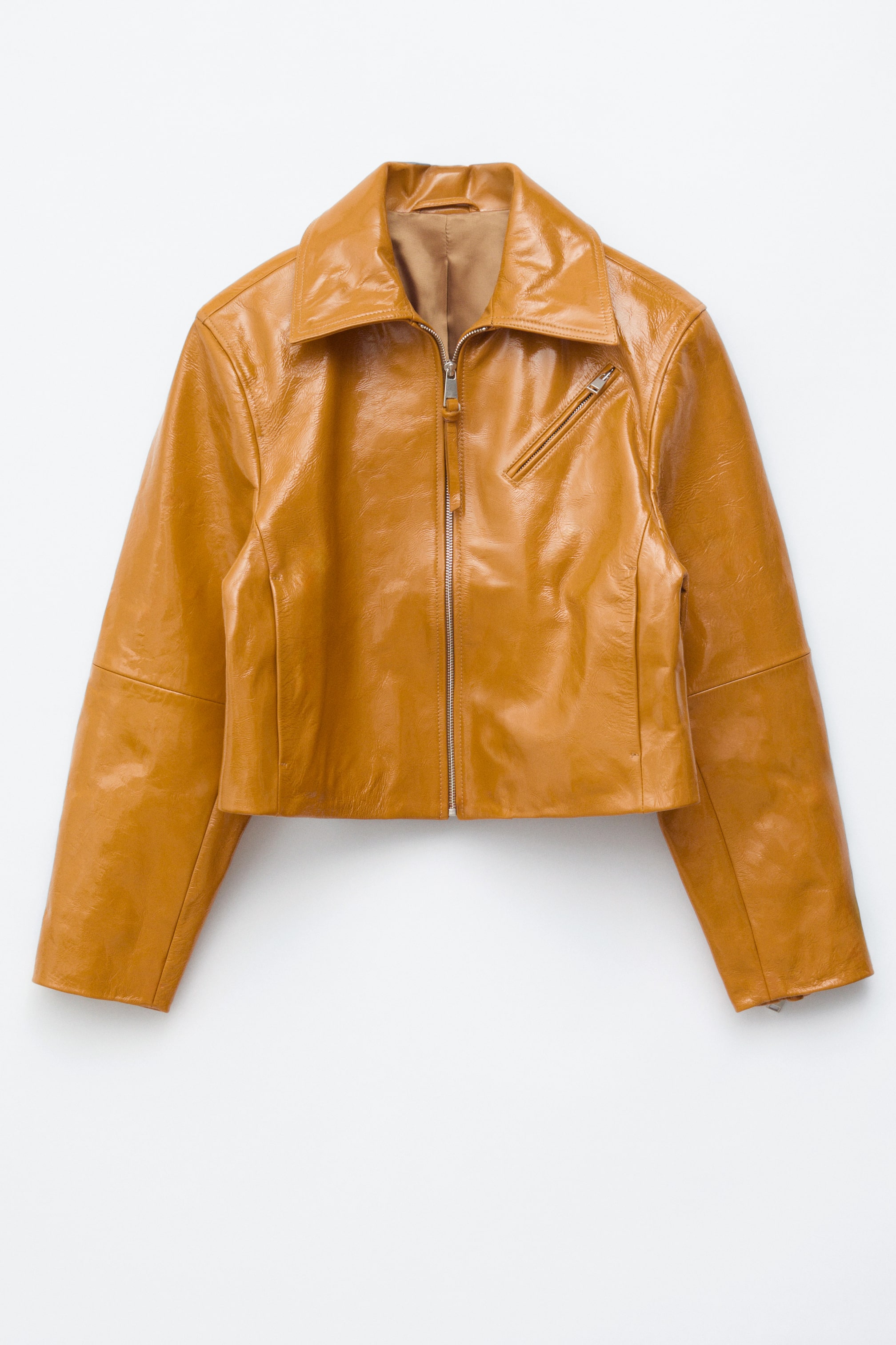 Grösseres Bild ansehen: Eine hellbraune Lacklederjacke wird von vorne präsentiert und besticht durch einen hochglänzenden Schimmer sowie eine kurze, kastige Silhouette. Sie ist mit einem klassischen Spitzkragen, einem silberfarbenen Reißverschluss über die gesamte Länge, schrägen Schnittlinien und einer schrägen Reißverschlusstasche auf der linken Brust gestaltet.