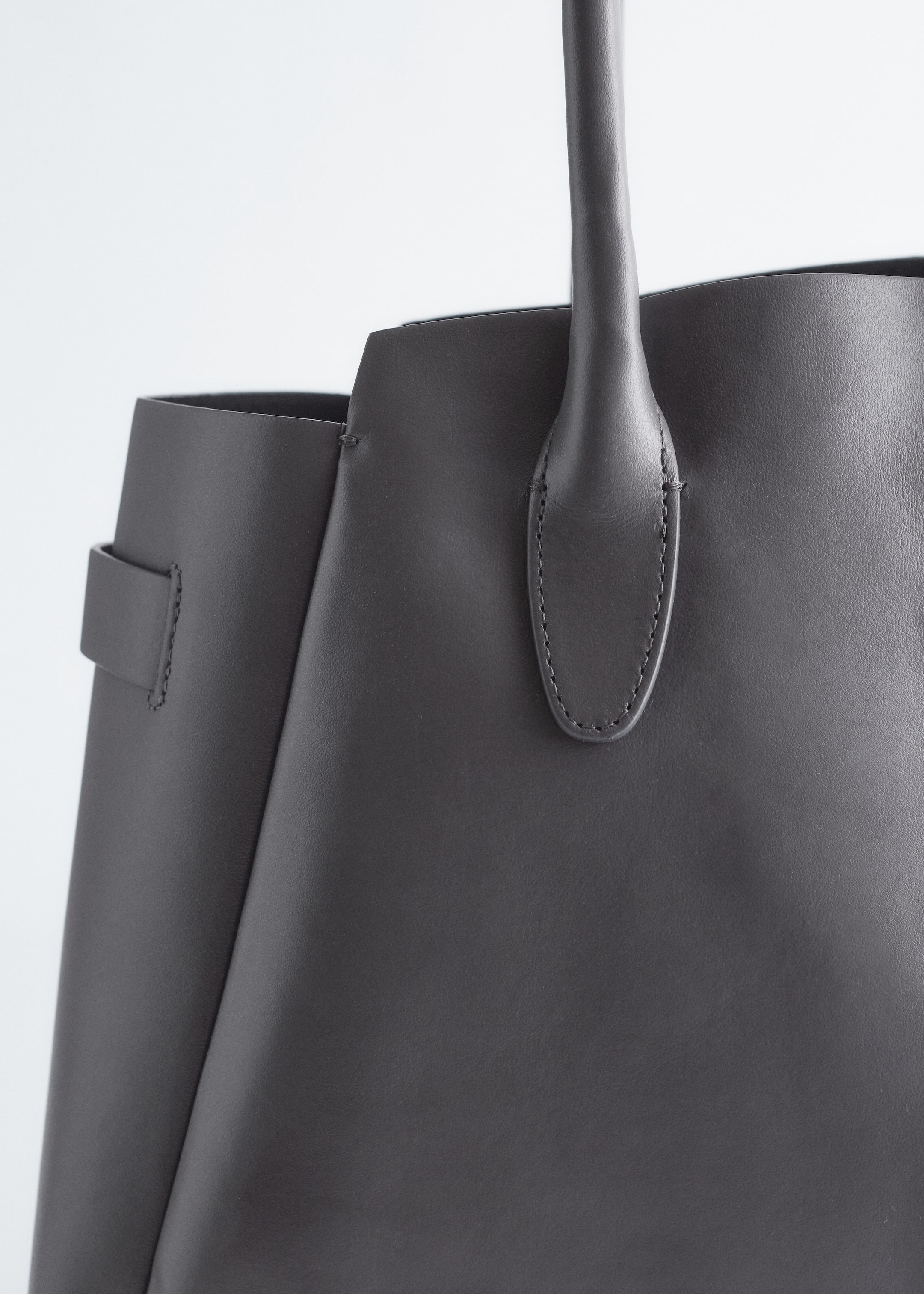 Visualizza immagine più grande: La borsa grigio scuro mostra una texture liscia sulla sua superficie anteriore, dove un punto di attacco del manico è rinforzato con cuciture visibili. Un pannello a soffietto laterale con una piccola linguetta cucita è anche visibile a sinistra.