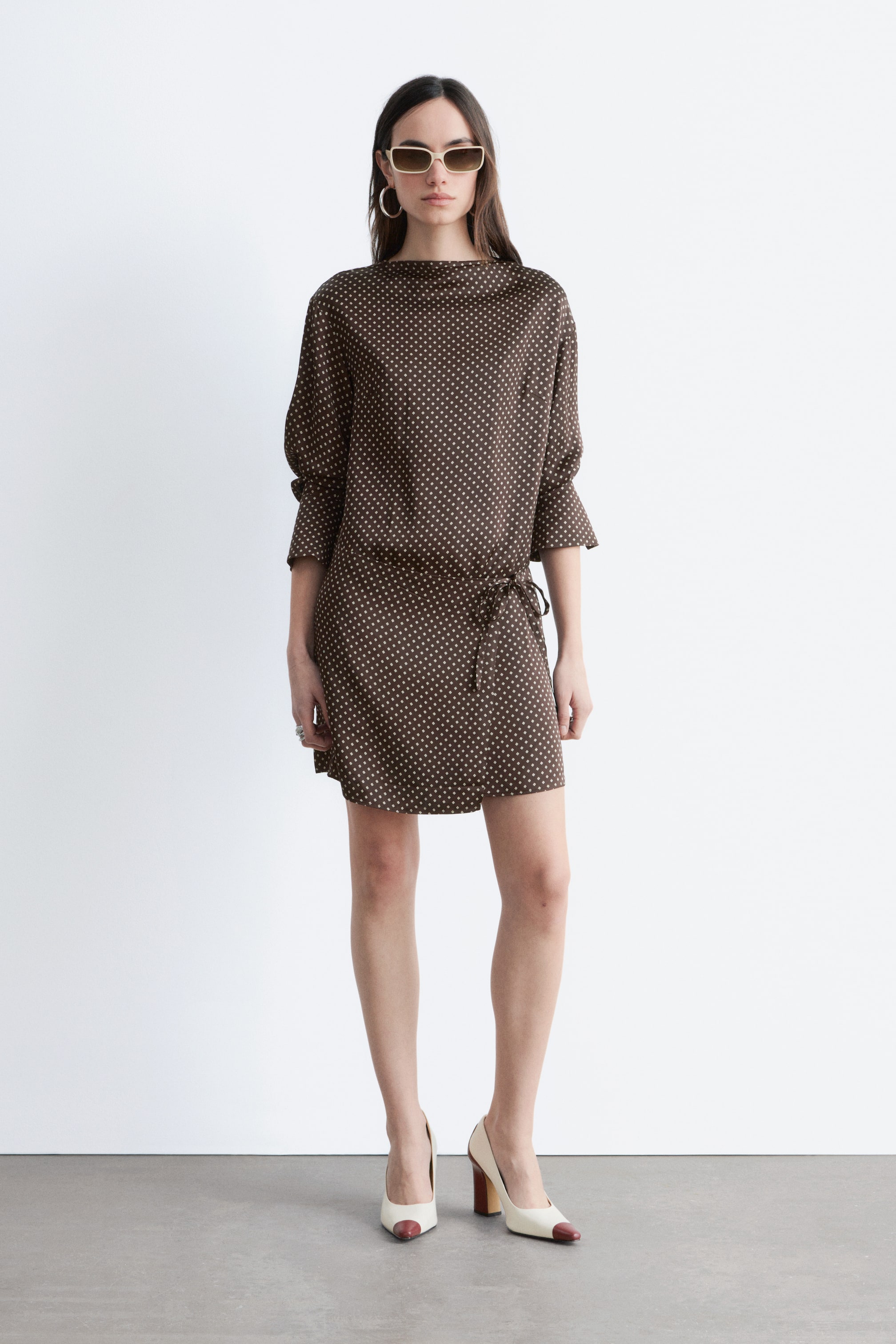 Grotere afbeelding bekijken: Een bruine jurk met witte stippen heeft een boothals en driekwart mouwen die bij de manchetten zijn geplooid. De mini-jurk is in de taille aangesnoerd met een zelfbindend trekkoord.