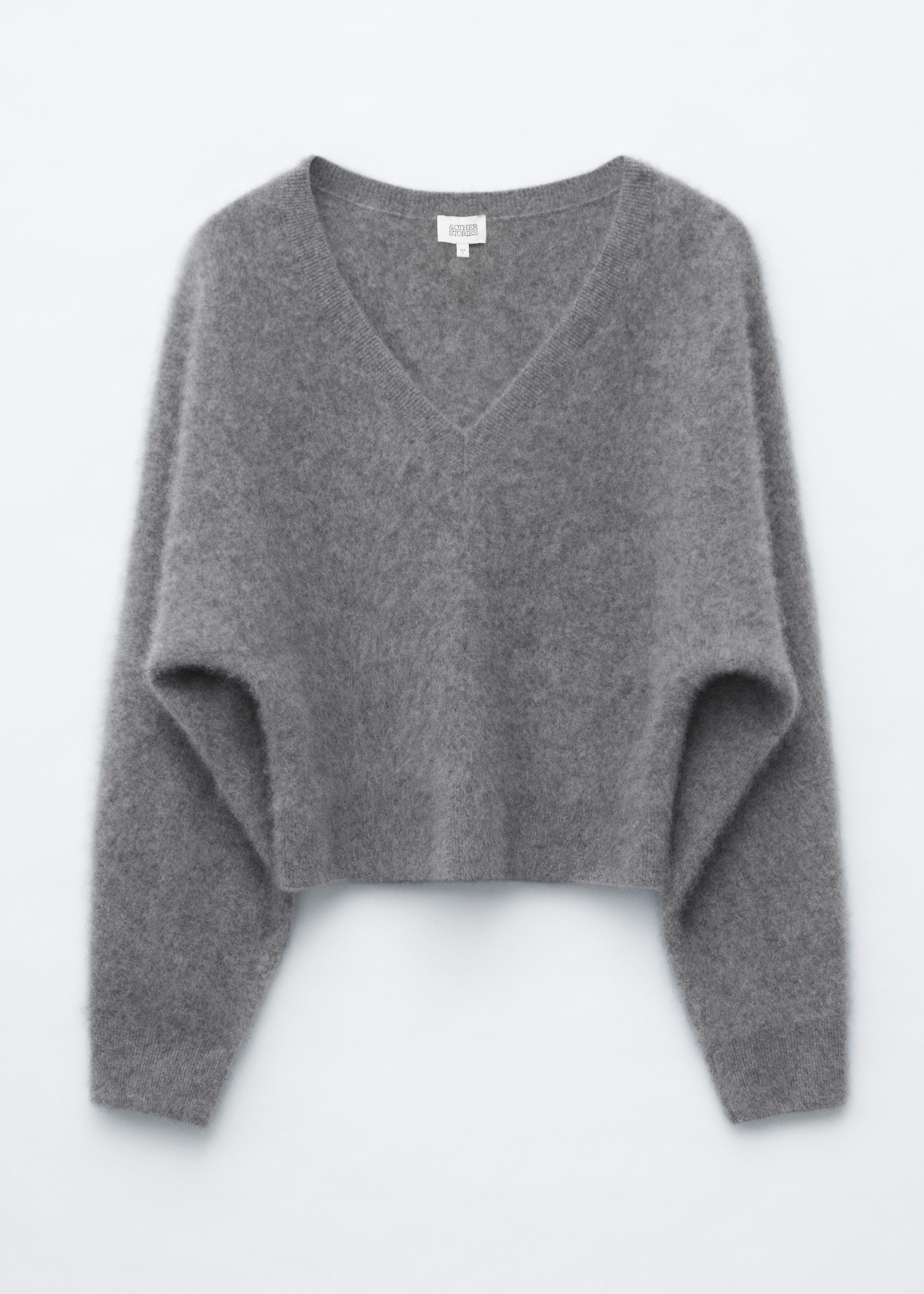 A grey brushed cashmere V-neck jumper is worn by the model and tucked into high-waisted grey trousers. The long-sleeved textured knit is styled with a silver pendant necklace resting over the neckline.