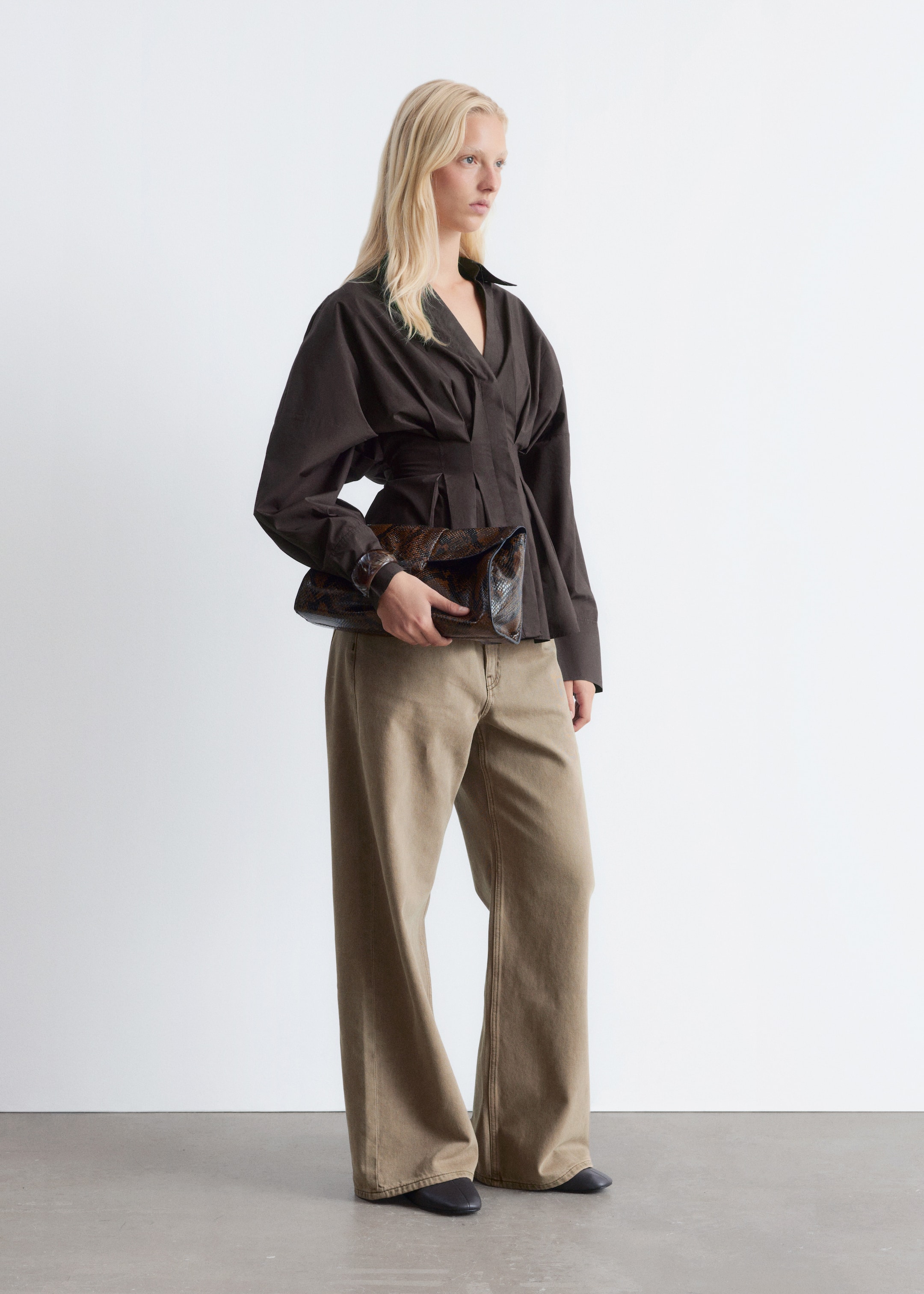 Agrandir l'image: Un haut marron foncé est présenté, avec un col en V, une taille structurée et plissée, et des manches volumineuses rassemblées aux poignets. Il est porté avec un pantalon kaki à jambes larges et taille haute, et un grand sac pochette à imprimé animal marron foncé et marron clair, avec un design plié, est tenu.