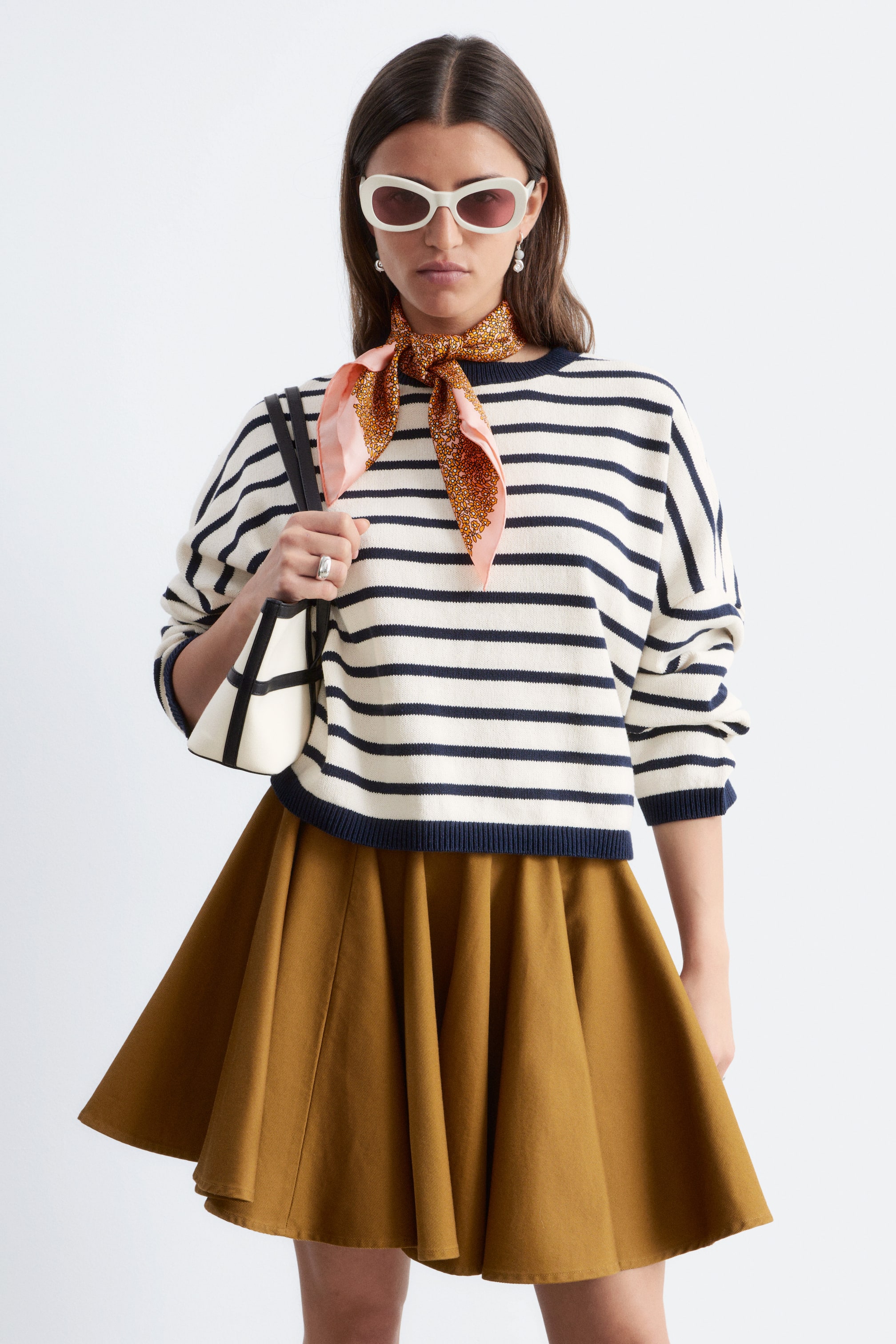 Visualizza immagine più grande: Il maglione presenta strisce orizzontali in bianco sporco e blu navy, rivolte in avanti. È progettato con un girocollo e polsini e orlo a coste in blu navy, mentre una sciarpa con motivo è legata intorno al collo e una gonna plissettata marrone è visibile sotto l'orlo.