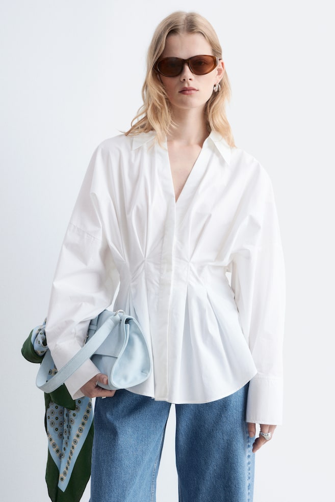 Ein weißes Hemd mit offenem V-Ausschnitt, taillierter Taille mit Faltendetails und voluminösen langen Ärmeln mit Knopfmanschetten. Das Model trägt das Hemd mit blauen Jeans und hält eine hellblaue Tasche sowie ein gemustertes Tuch. - 1