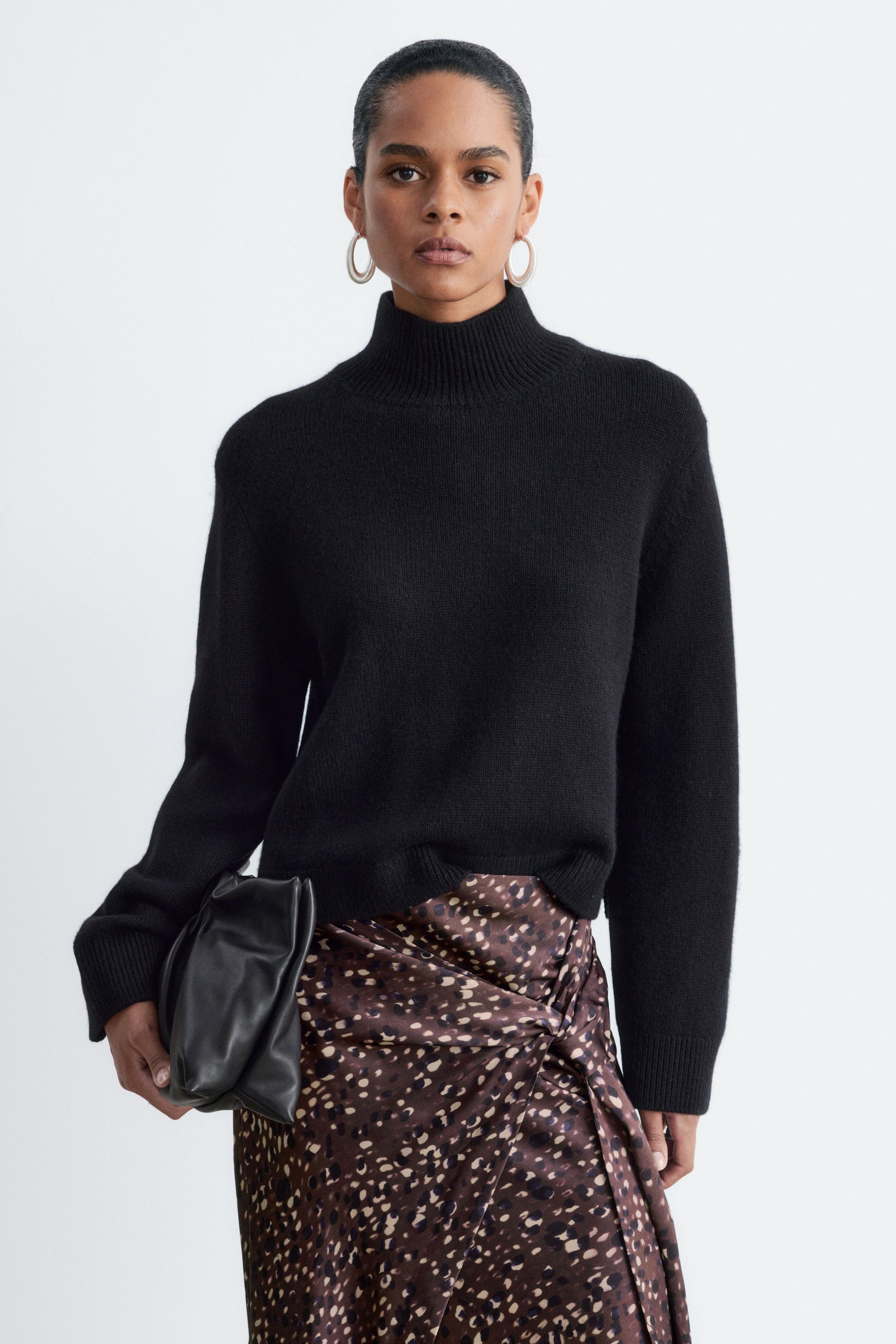 Visualizza immagine più grande: Un maglione nero in misto lana e cashmere con collo alla coreana, caratterizzato da colletto e polsini a coste, è indossato dalla modella. Il maglione è portato leggermente infilato in una gonna a portafoglio stampata, mentre la modella tiene in mano una pochette in pelle nera.
