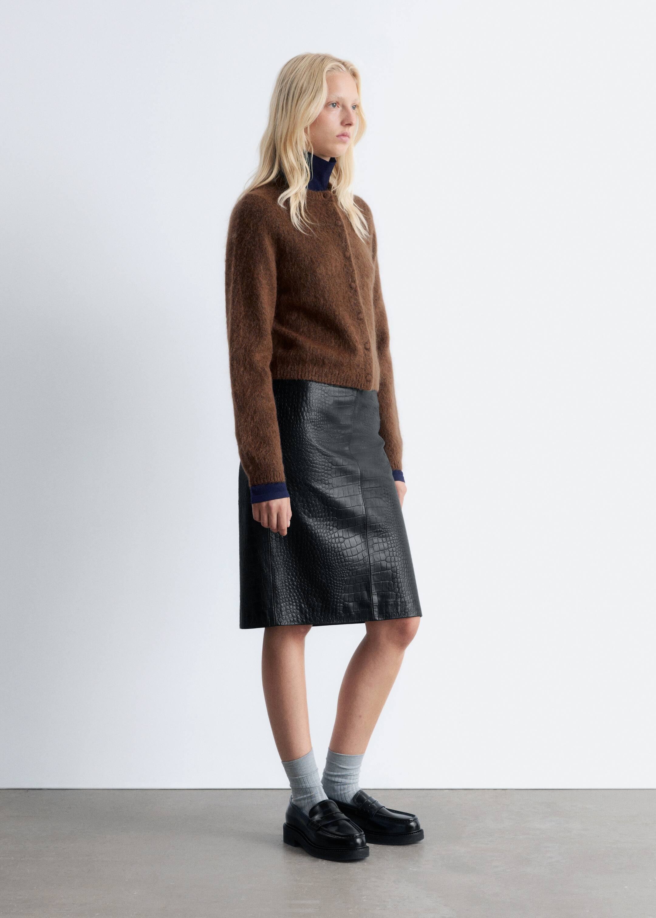Agrandir l'image: Un cardigan marron duveteux, avec un col rond et une fermeture à boutons sur le devant, est porté par-dessus un col roulé bleu marine. Une jupe midi noire embossée façon crocodile avec une silhouette droite est présentée, accompagnée de mocassins noirs épais et de chaussettes côtelées gris clair.