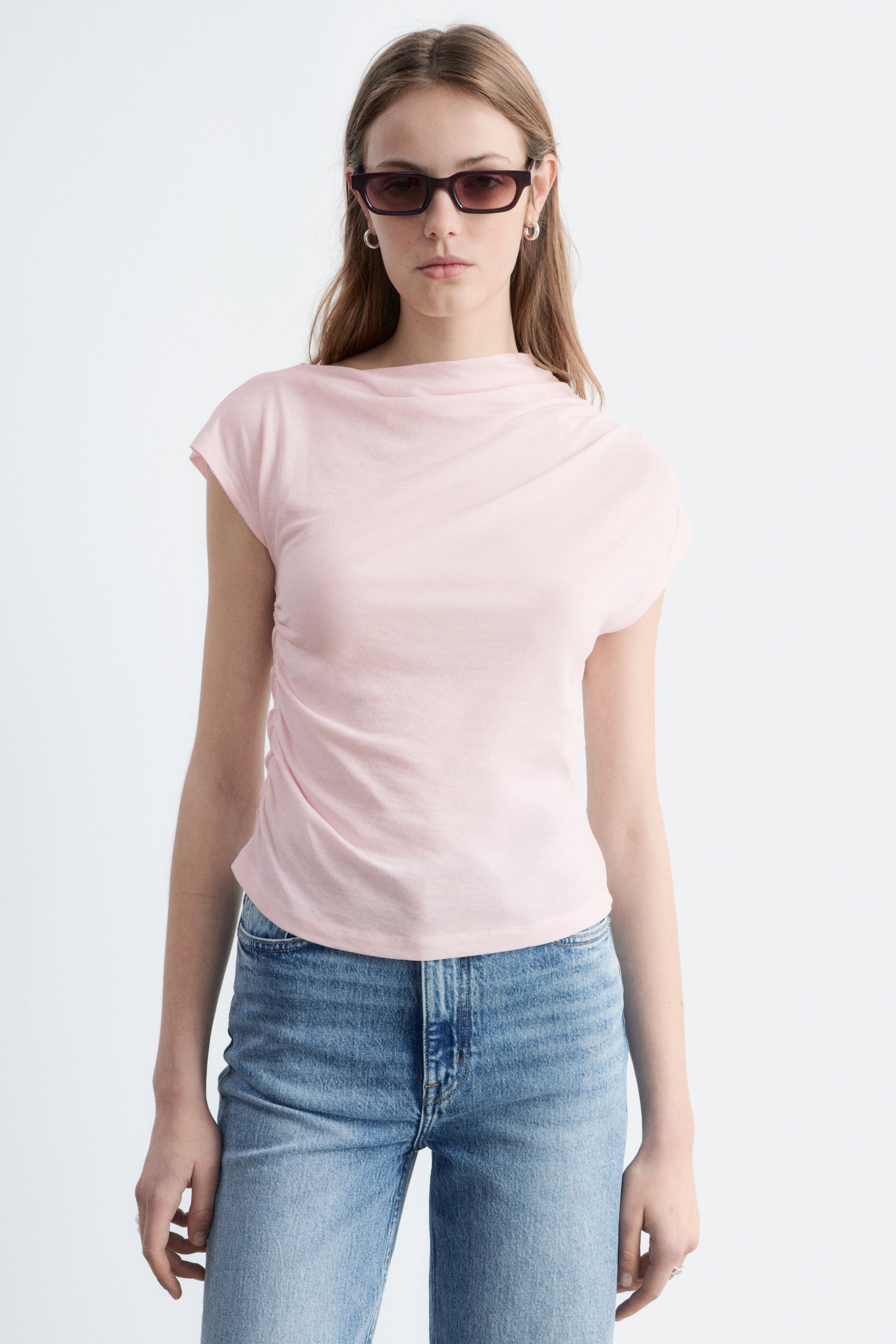 Größeres Bild anzeigen: Ein hellrosa Oberteil mit Kappärmeln und gerafften Nähten wird von dem Model getragen. Die Raffungen erzeugen einen drapierten Effekt, der vom Ausschnitt ausgeht und sich entlang beider Seiten des Oberkörpers erstreckt. Das Oberteil hat eine schmale Passform und wird mit blauen Jeans kombiniert.