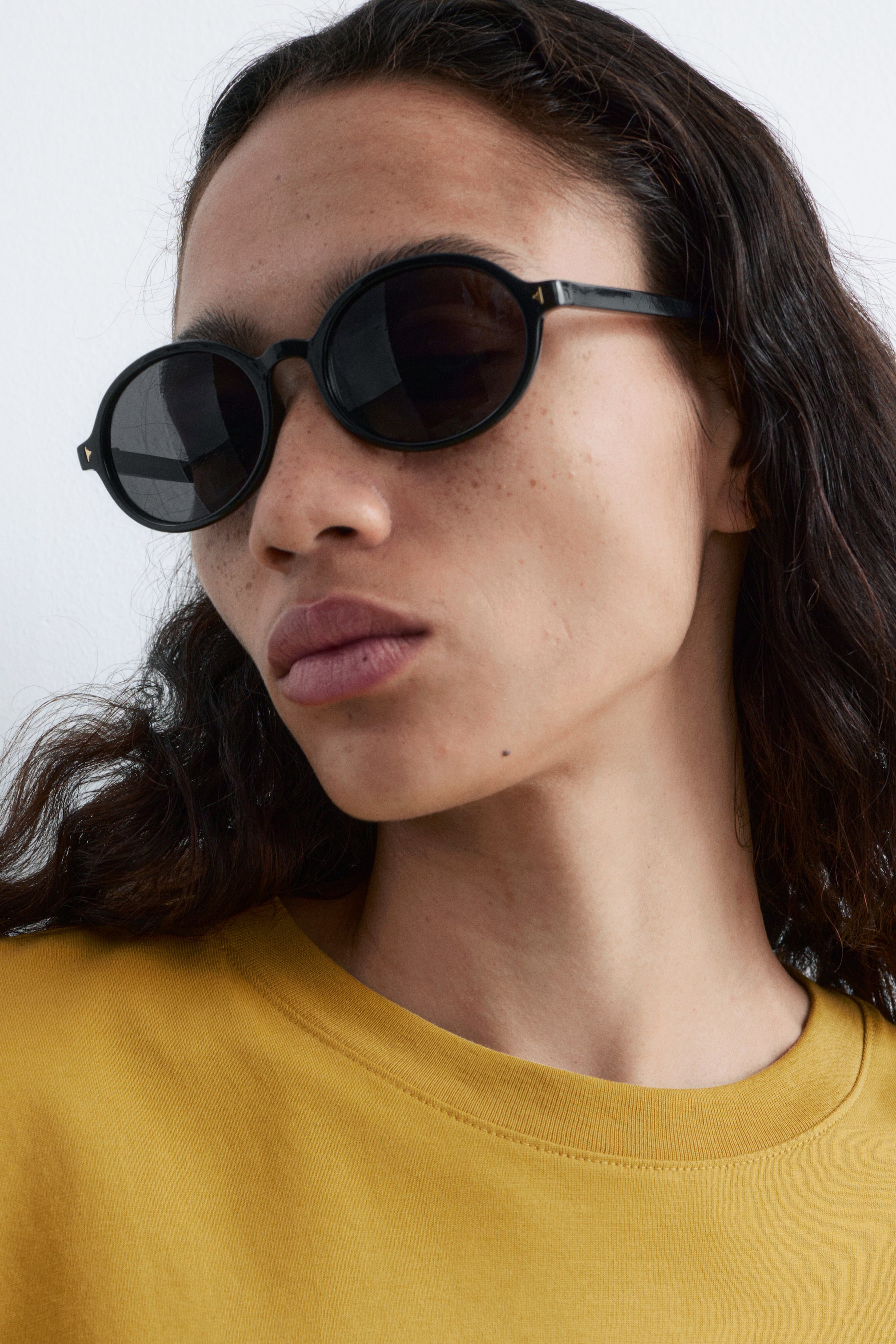 View larger image: Black sunglasses are being worn, angled slightly to the side, featuring round frames and dark, tinted lenses. A small golden triangular detail is visible on the outer edge of each frame near the temple.