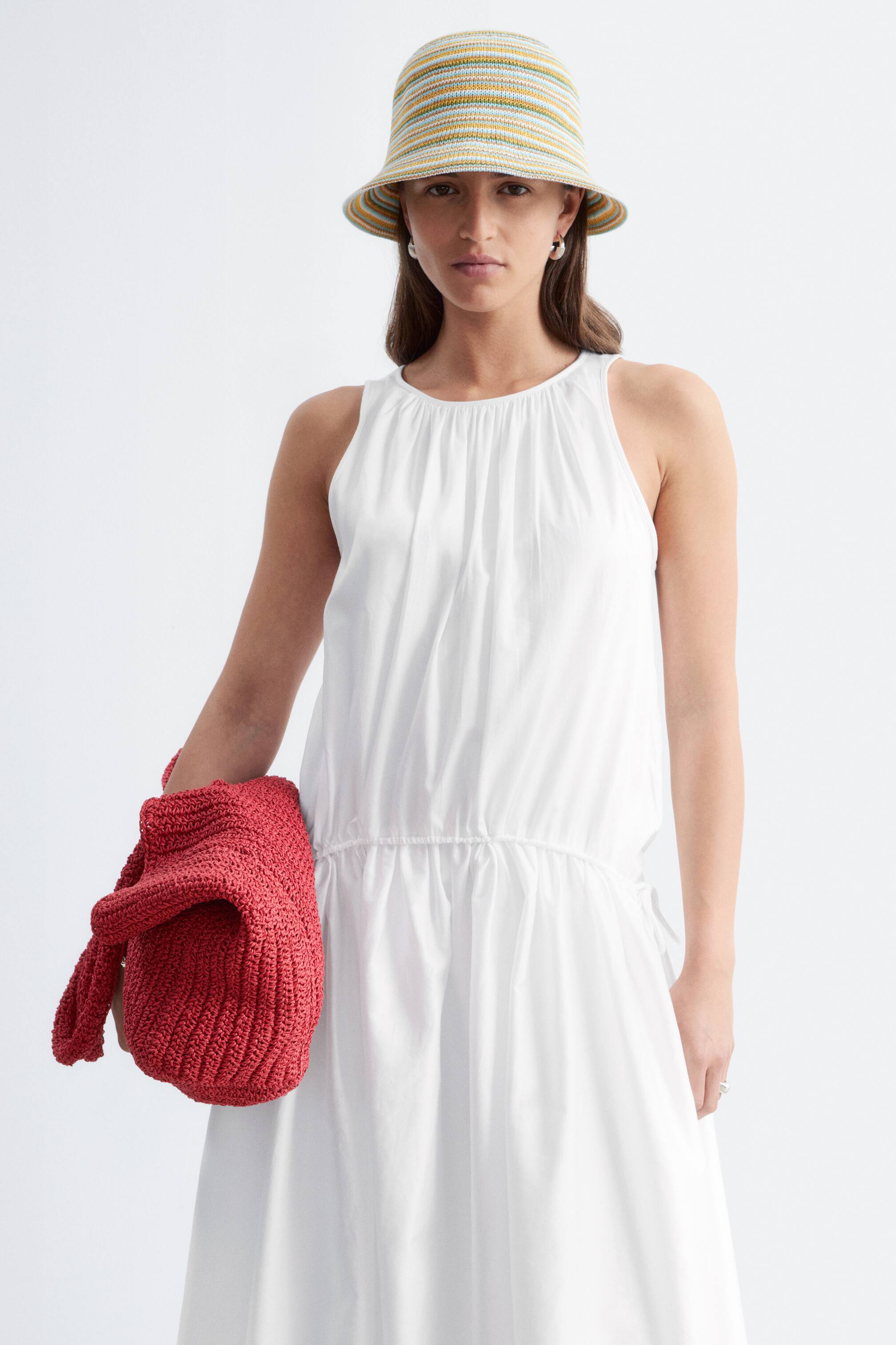 Agrandir l'image: La mannequin porte une robe blanche sans manches avec des fronces à l'encolure et une taille à cordon de serrage. Elle arbore également un bob rayé et tient un sac cabas en paille tressée rouge dans sa main gauche.