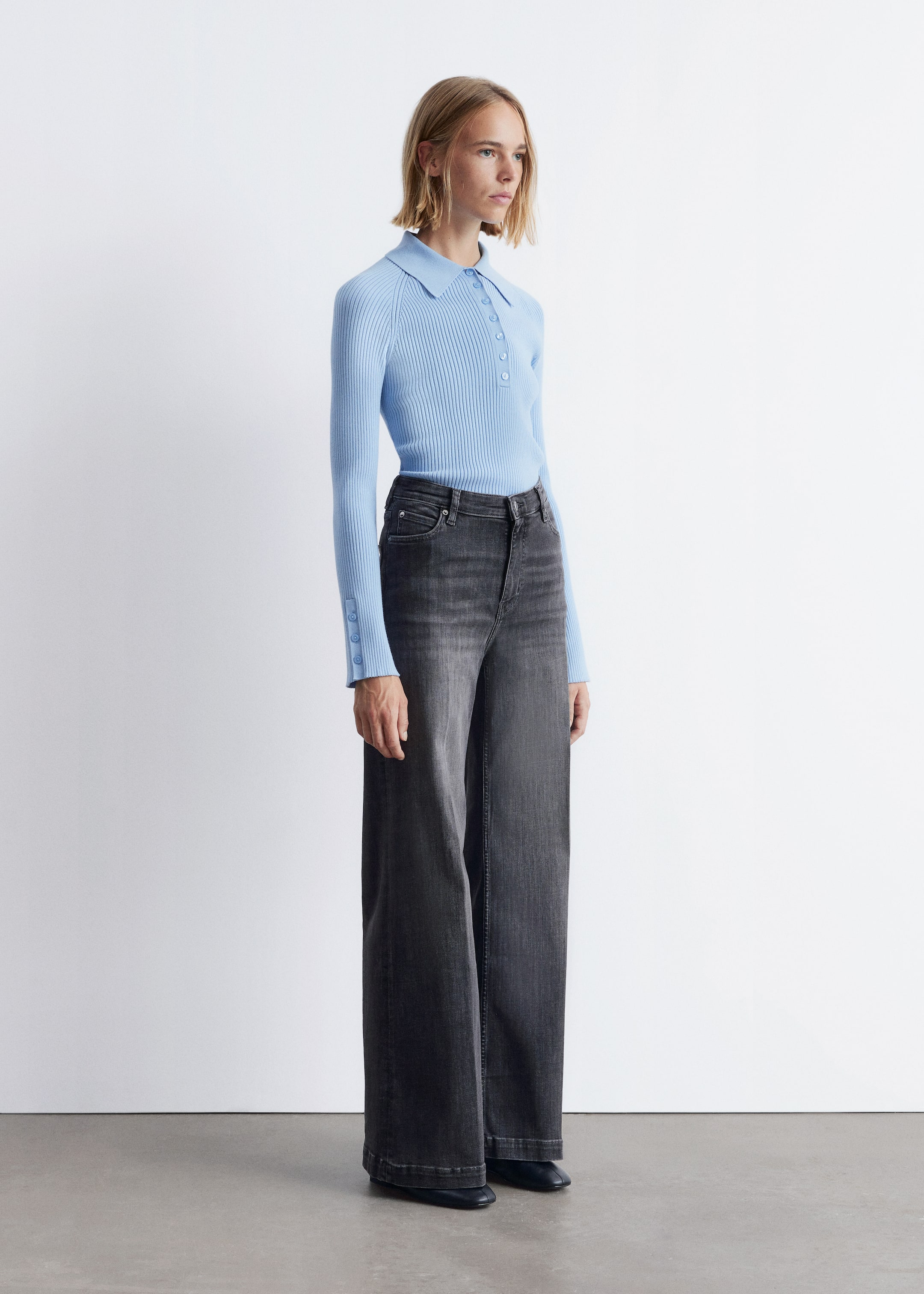 Visualizza immagine più grande: Un top polo a maglia a coste azzurro chiaro, con un colletto classico e una chiusura a tre bottoni, è indossato. Ha maniche lunghe con polsini a tre bottoni ed è abbinato a jeans a vita alta, grigio scuro, a gamba larga, che mostrano le silhouette distintive da una vista laterale.