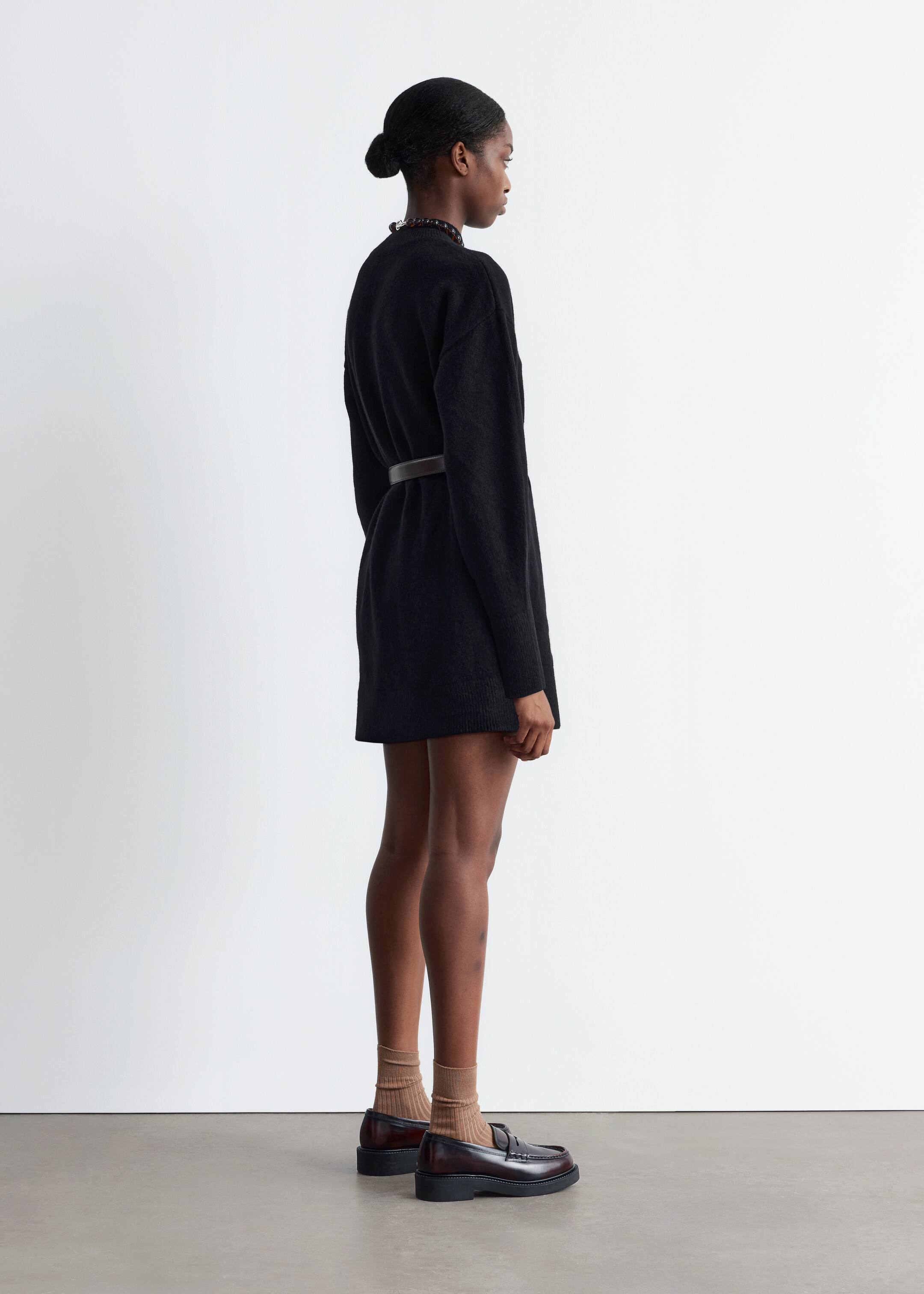 Größeres Bild anzeigen: Ein schwarzes, locker sitzendes Mini-Strickkleid mit einem Stehkragen wird von dem Model getragen. Ein tonaler Schnallengürtel betont die Taille des Models.