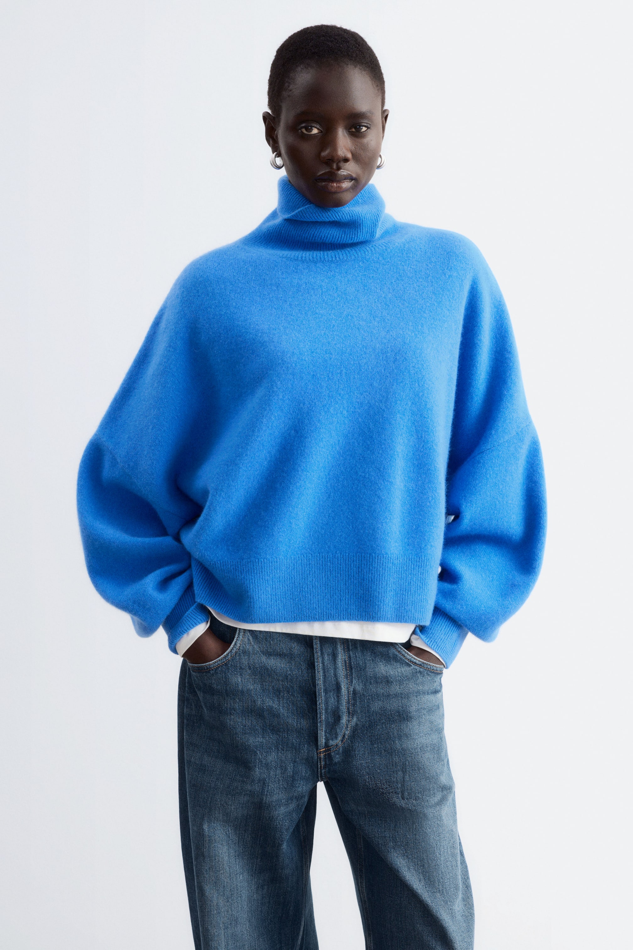 View larger image: An oversized, bright blue knitted jumper featuring a folded rib turtleneck, dropped shoulders, and voluminous sleeves is being worn by the model. The jumper is layered over a white top and paired with blue jeans, with the model placing their hands inside the front pockets.