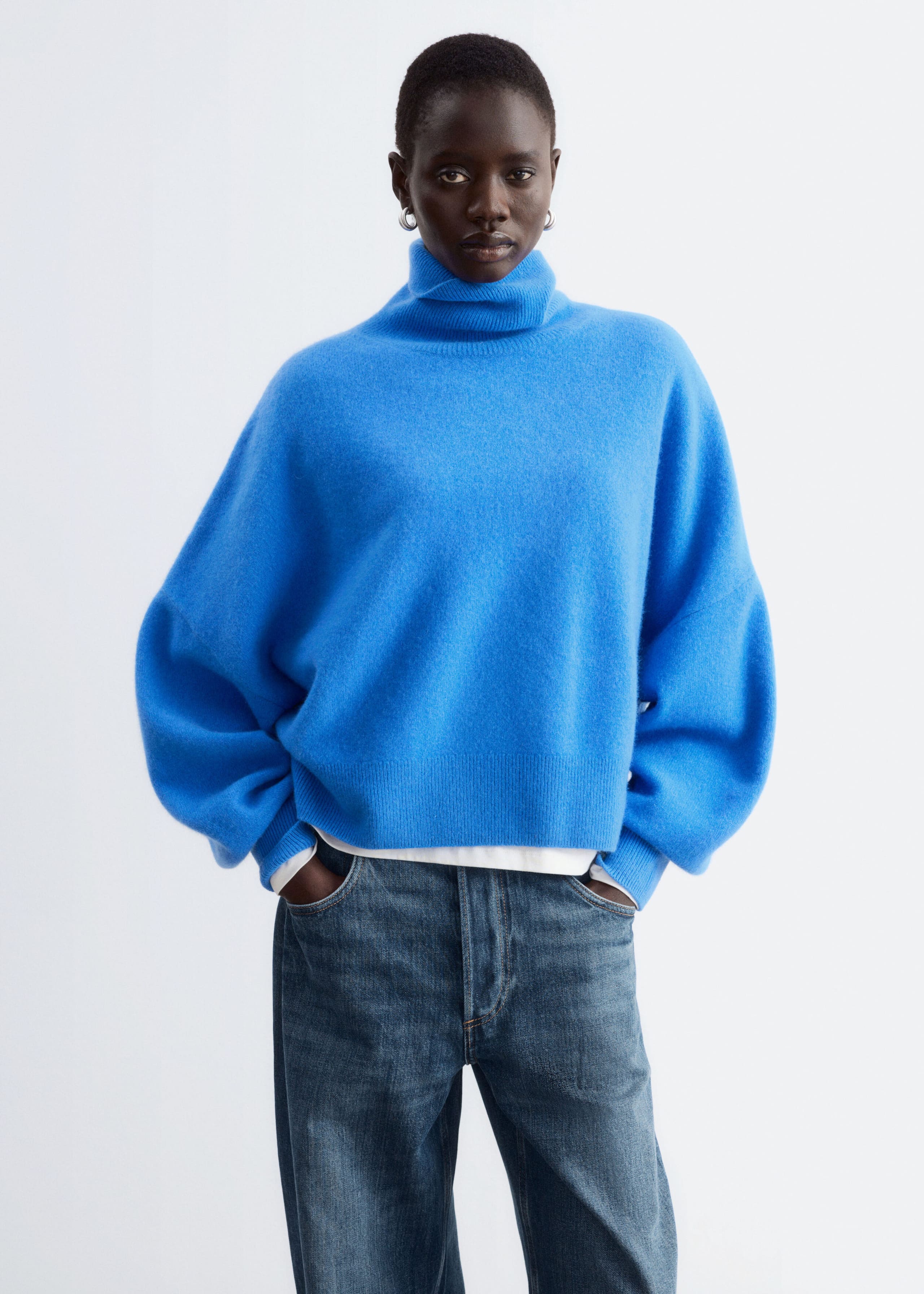 An oversized, bright blue knitted jumper featuring a folded rib turtleneck, dropped shoulders, and voluminous sleeves is being worn by the model. The jumper is layered over a white top and paired with blue jeans, with the model placing their hands inside the front pockets.