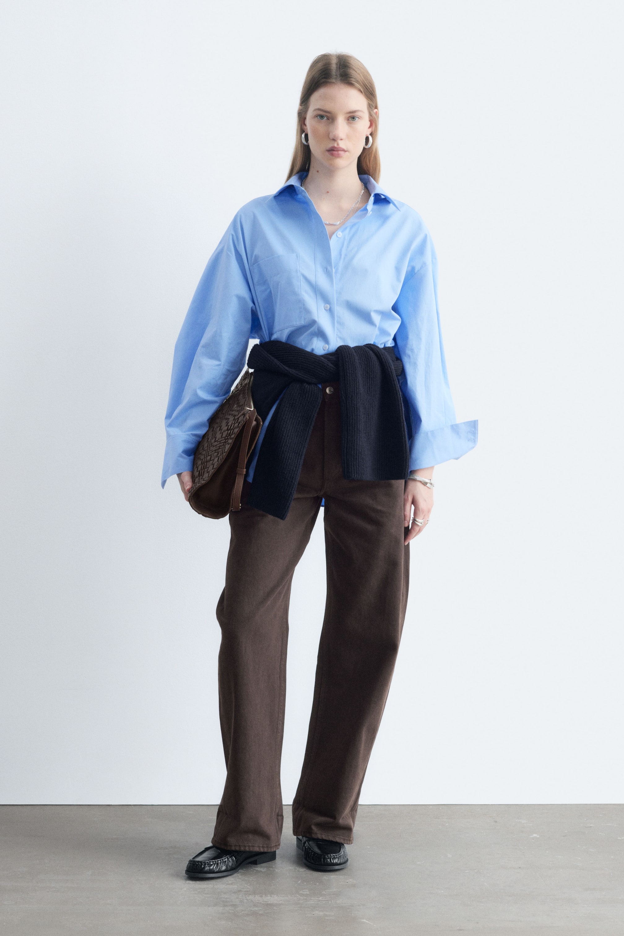 Grotere afbeelding bekijken: Bruine jeans, met een oversized pasvorm en extra lange, barrel-vormige pijpen, worden gedragen door het model. De mid-waist jeans zijn gestyled met een lichtblauw shirt, en een donkere trui is rond de taille geknoopt, terwijl het model een bruine tas draagt.