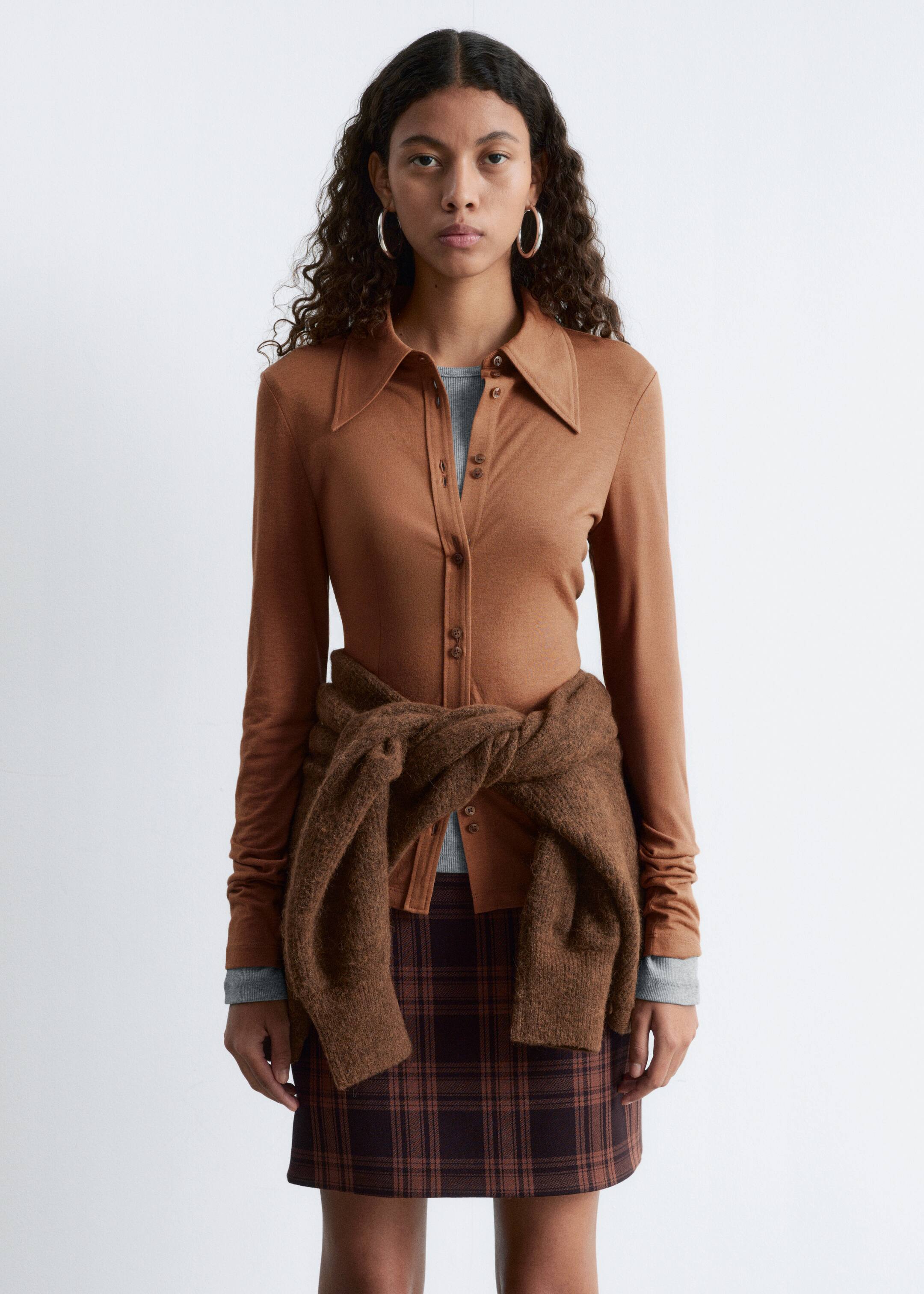 Agrandir l'image: La chemise marron à manches longues est orientée vers l'avant, avec un col pointu et une patte de boutonnage à l'avant avec des boutons visibles. Elle est superposée à un haut gris à manches longues, avec un pull marron en tricot noué autour de la taille au-dessus de la chemise.