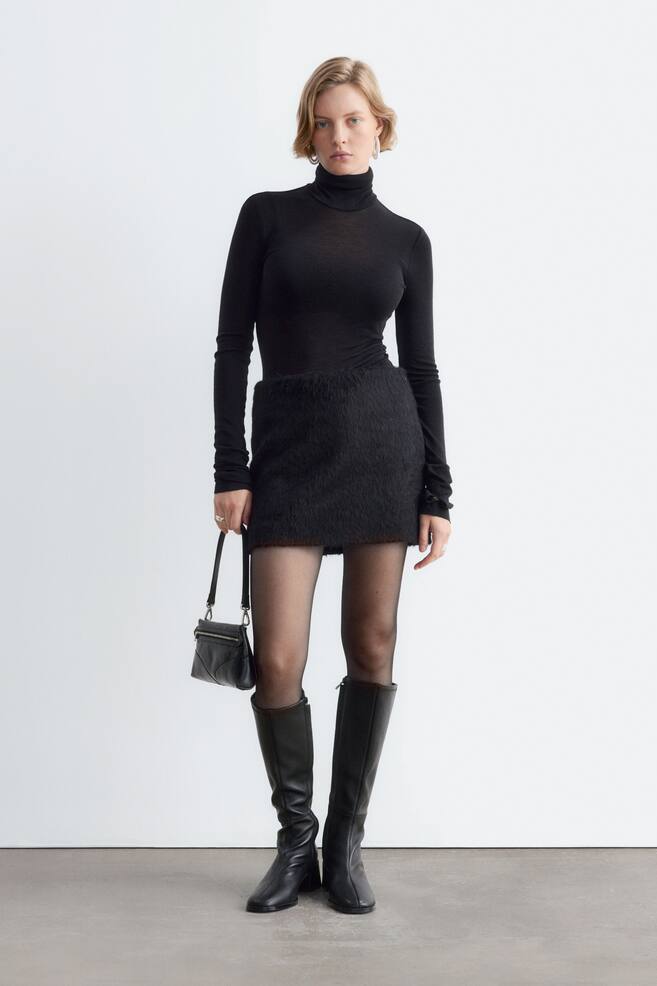 The model is wearing a black textured mini skirt, styled with a fitted black roll-neck top, sheer black tights, and knee-high black leather boots. She is standing and carrying a small black shoulder bag. - 1