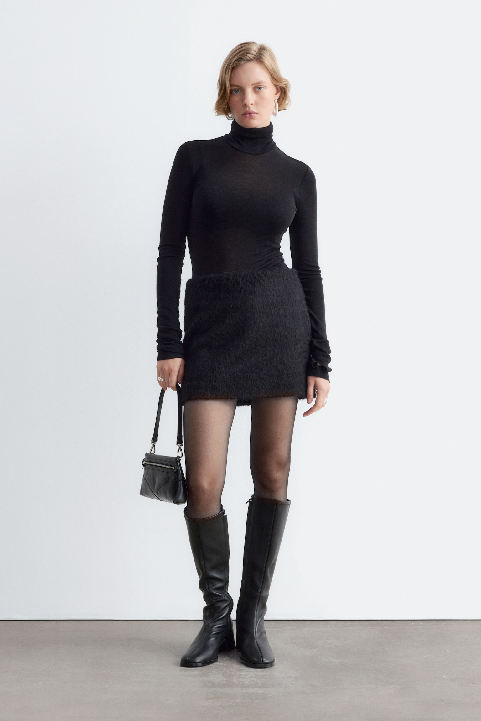 The model is wearing a black textured mini skirt, styled with a fitted black roll-neck top, sheer black tights, and knee-high black leather boots. She is standing and carrying a small black shoulder bag. - 1