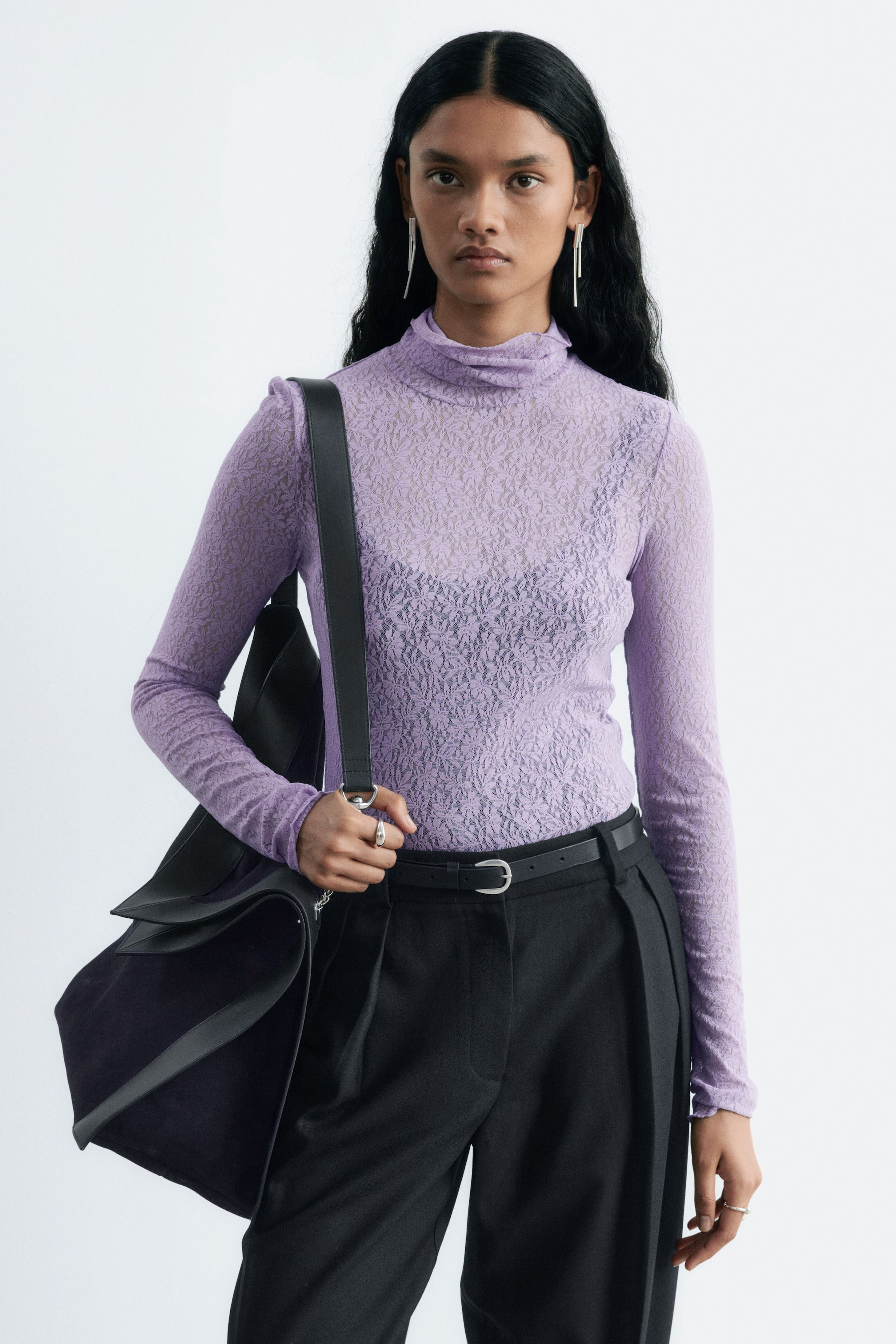 Grotere afbeelding bekijken: Een lavendelkleurige kanten jersey top met een textuurpatroon, col en lange mouwen wordt gedragen door het model. De top is nauwsluitend en laat een donker onderhemd zien onder de transparante stof, en aan de mouwen zijn duimgaten zichtbaar.