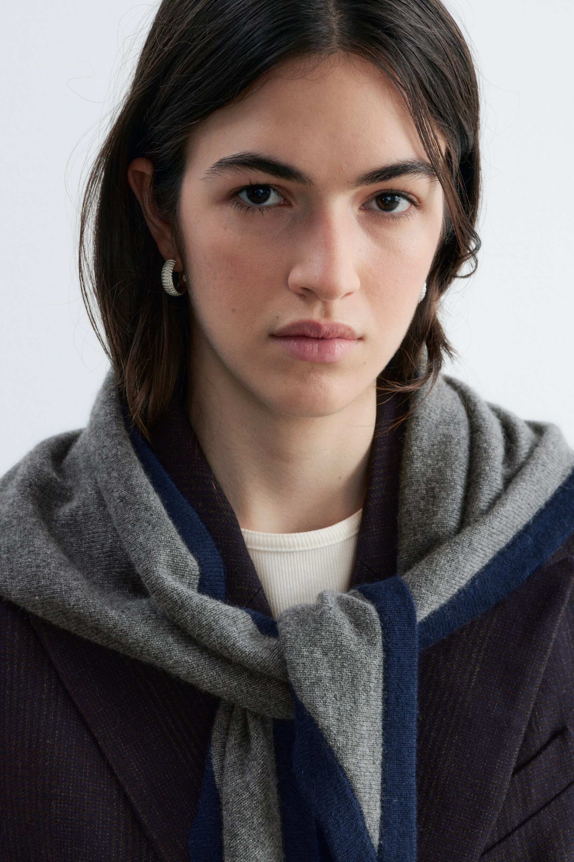 View larger image: A silk-cashmere triangle scarf in grey with a contrasting dark blue border is worn by the model, draped over the shoulders and loosely knotted at the front. The scarf is layered over a dark pinstripe blazer and a white ribbed top.