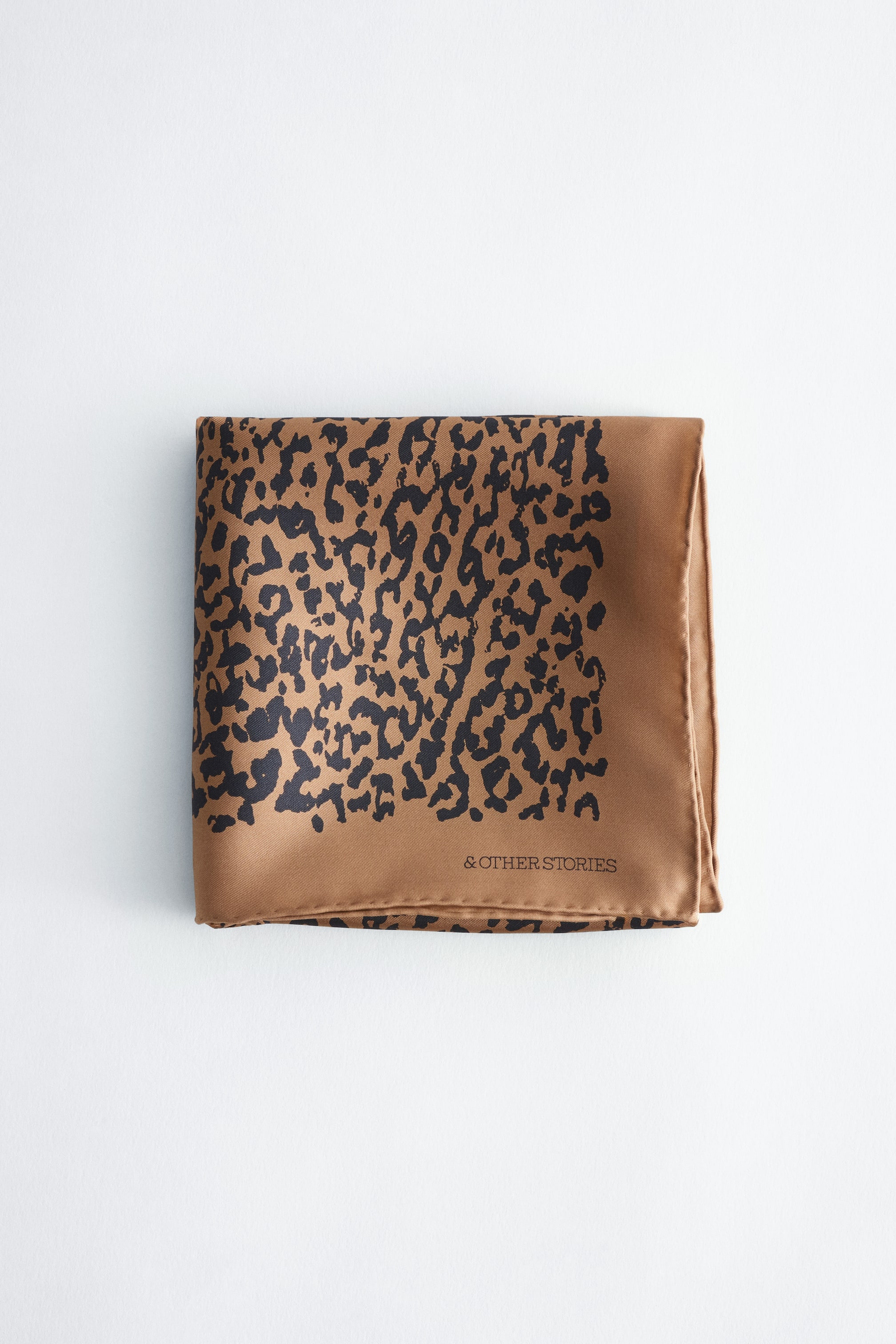 Vezi imaginea mai mare: O eșarfă din mătase imprimată, împăturită în formă de pătrat, afișează un model negru cu imprimeu leopard pe un fundal bej-închis. Eșarfa are o bordură uni bej-închis de-a lungul marginilor de jos și din dreapta, iar numele brandului „& OTHER STORIES” este imprimat cu negru în colțul din dreapta jos.