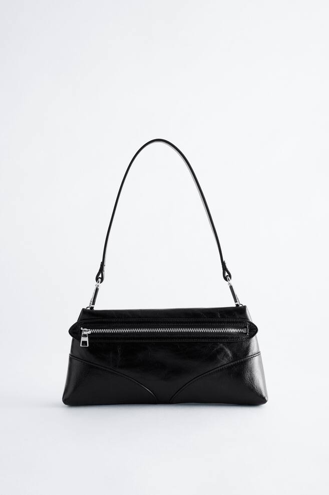 Un sac bandouliÃ¨re noir est prÃ©sentÃ© de face, avec une forme rectangulaire structurÃ©e, une finition lÃ©gÃ¨rement brillante et un matelassage cousu incurvÃ© sur la partie infÃ©rieure. Le sac prÃ©sente une poche frontale horizontale avec une fermeture Ã©clair en mÃ©tal argentÃ© et est suspendu par une seule bandouliÃ¨re fine, fixÃ©e Ã  lâaide de ferrures ton argent. - 1