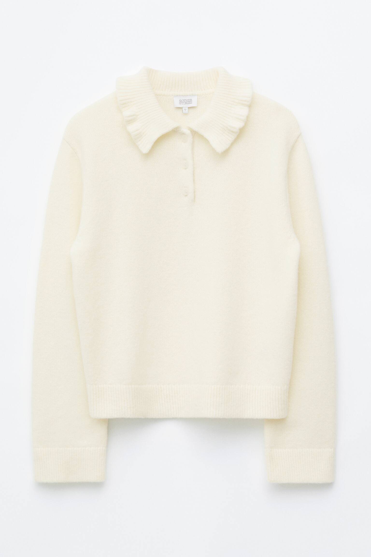 En krÃ¤mfÃ¤rgad jumper med volangkrage Ã¤r vÃ¤nd framÃ¥t och visar en mjuk stickad design med en volangprydd polokrage och en kort knappslÃ¥ med tre knappar. Den lÃ¥ngÃ¤rmade trÃ¶jan visar en ribbstickad nederkant och matchande ribbstickade Ã¤rmslut.