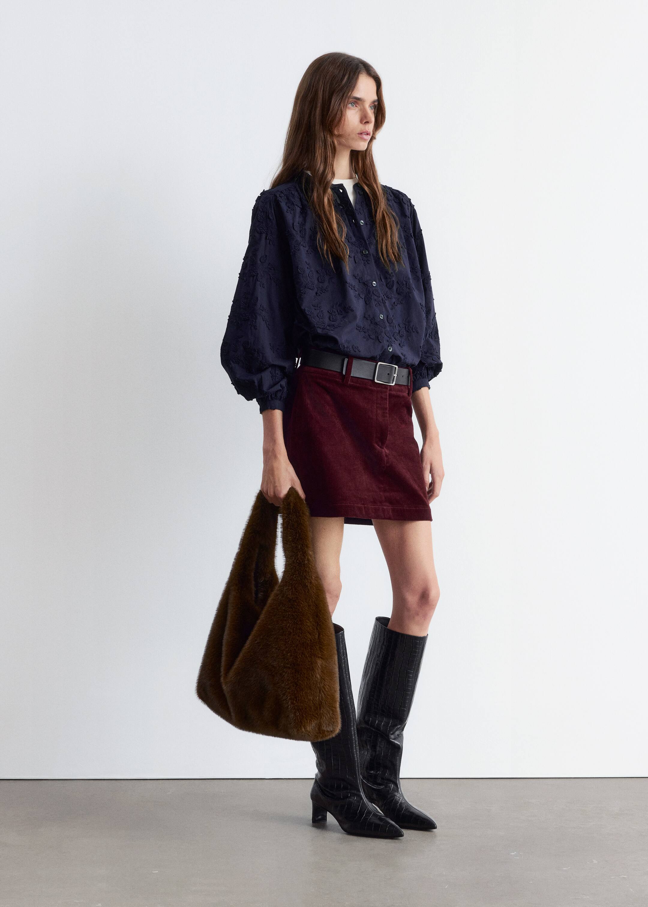Agrandir l'image: Une blouse bleu marine à boutons sur le devant avec un motif floral texturé et des manches volumineuses avec des poignets froncés est présentée, rentrée dans une mini-jupe en velours côtelé bordeaux. Une ceinture noire avec une boucle en argent serre la jupe, et des bottes noires embossées façon crocodile, montant jusqu'aux genoux, avec des bouts pointus et des talons blocs sont portées. Un sac hobo en fausse fourrure marron est tenu.