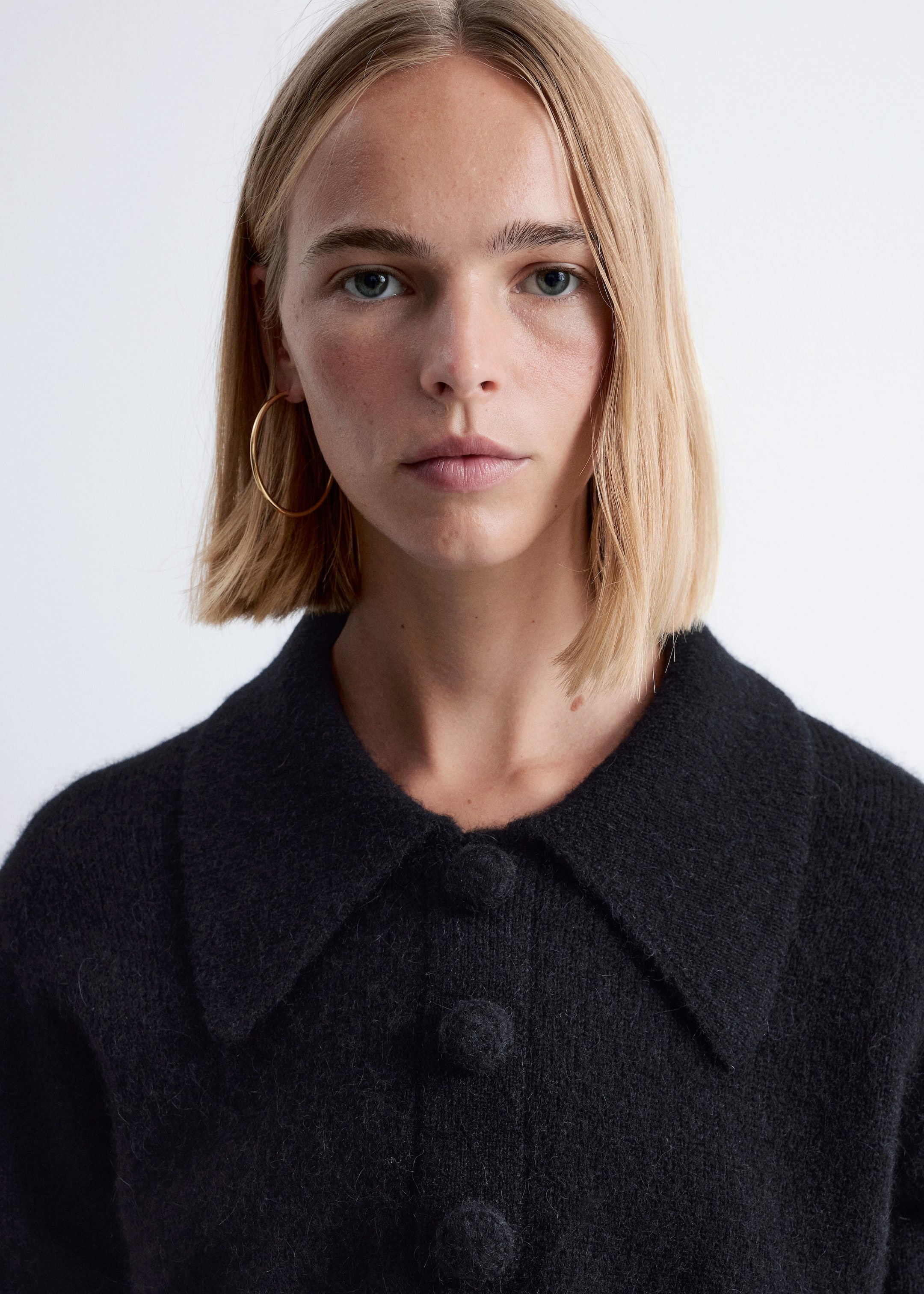 Agrandir l'image: Un cardigan en tricot brossé noir est porté, avec un col pointu classique. Trois grands boutons ronds recouverts de tissu ferment le devant du vêtement.