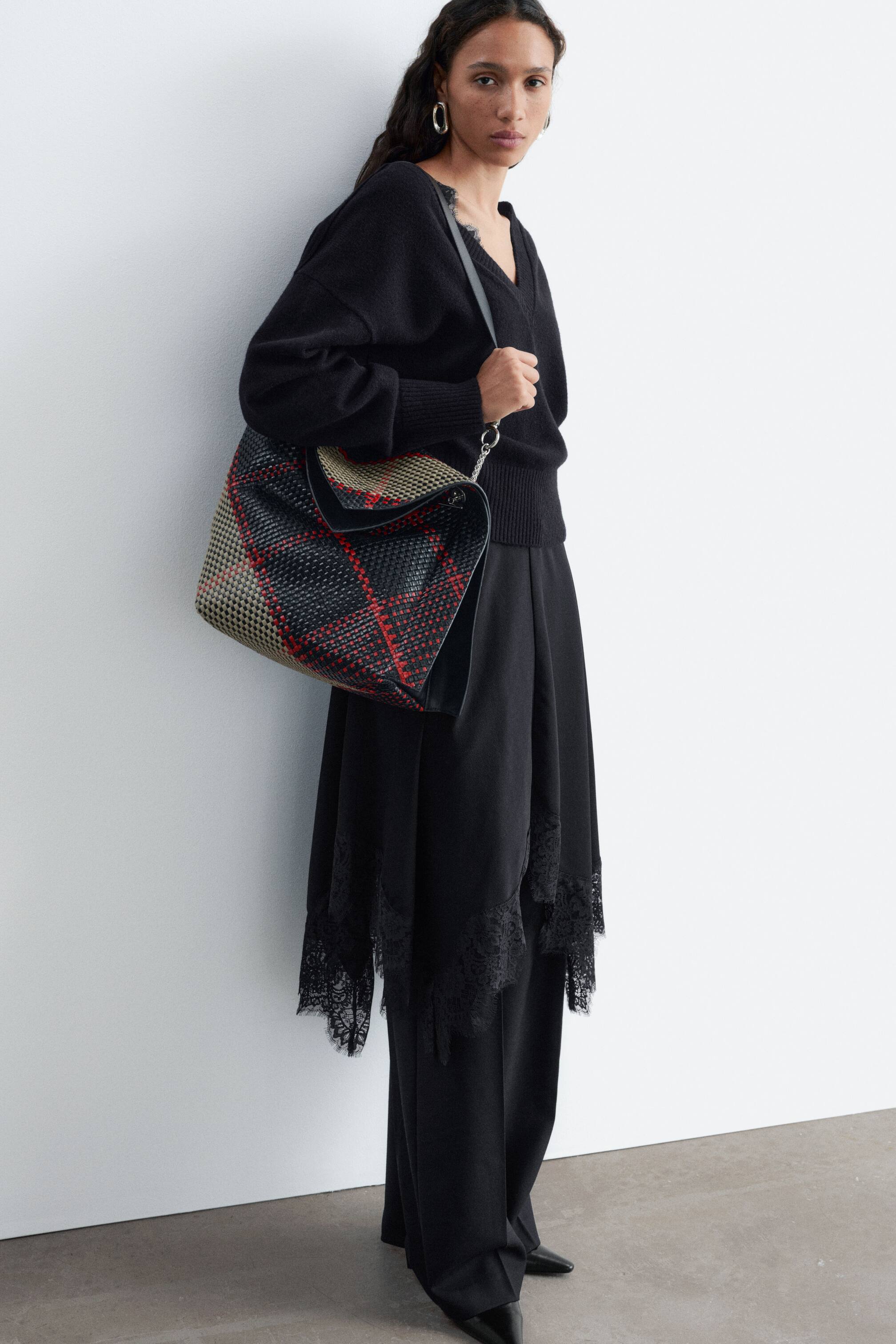 Grösseres Bild ansehen: Eine geflochtene Ledertasche mit schwarz-, rot- und beigefarbenem Karomuster wird von dem Model über der Schulter getragen, das den Trageriemen in der Hand hält. Die unstrukturierte Tasche verfügt über eine weiche, lässig fallende Silhouette mit geschwungener Öffnung und ist zu einem komplett schwarzen Strick- und Spitzenoutfit gestylt.