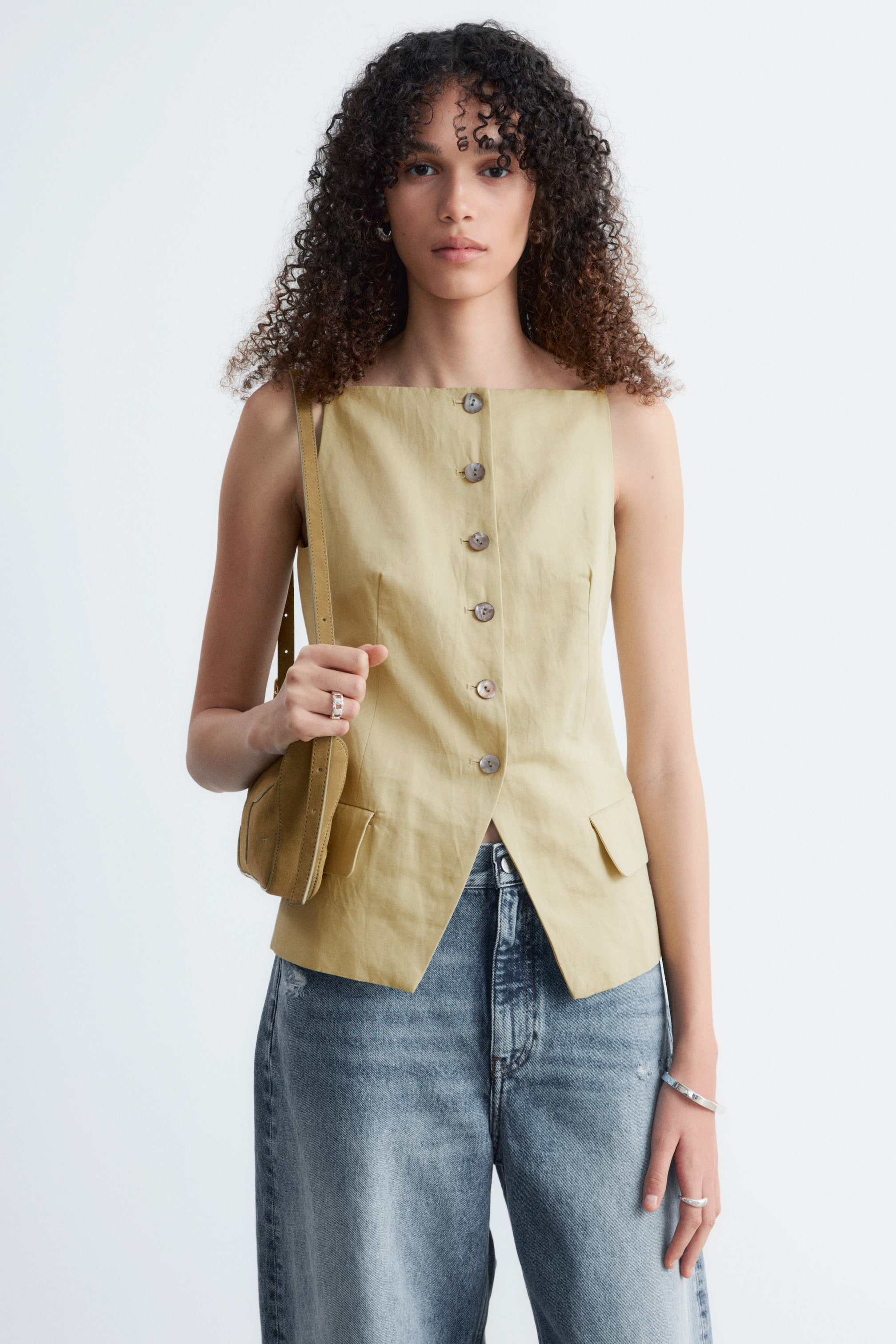 Visualizza immagine più grande: Un gilet senza maniche di colore beige chiaro presenta uno scollo quadrato e una chiusura frontale completa con bottoni rotondi. Sono visibili due tasche frontali con patta, e un orlo asimmetrico forma uno spacco frontale, posandosi sopra jeans blu.