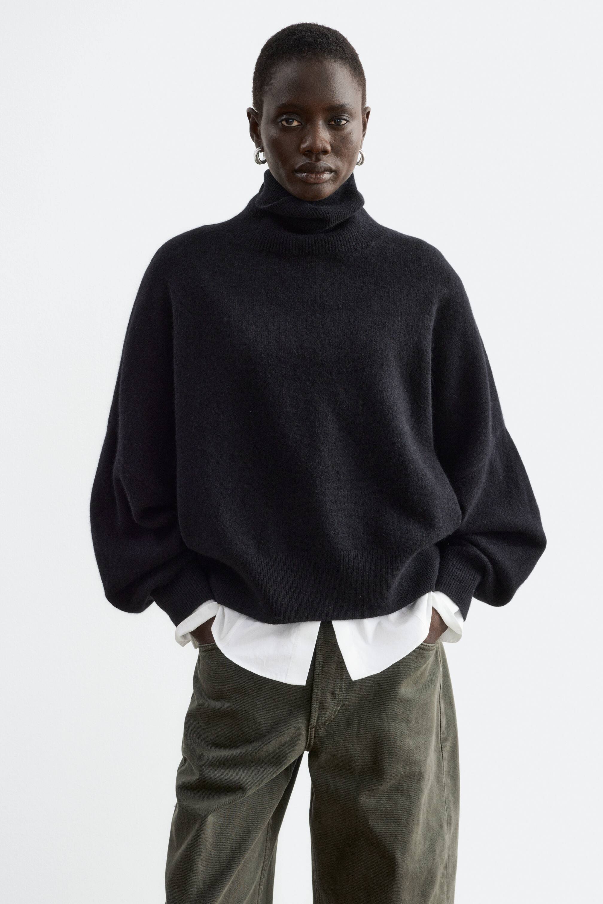 View larger image: An oversized black jumper featuring a folded rib turtleneck and dropped armholes is being worn by the model. The jumper is layered over a white collared shirt and styled with olive green trousers, with the model resting their hands in the pockets.