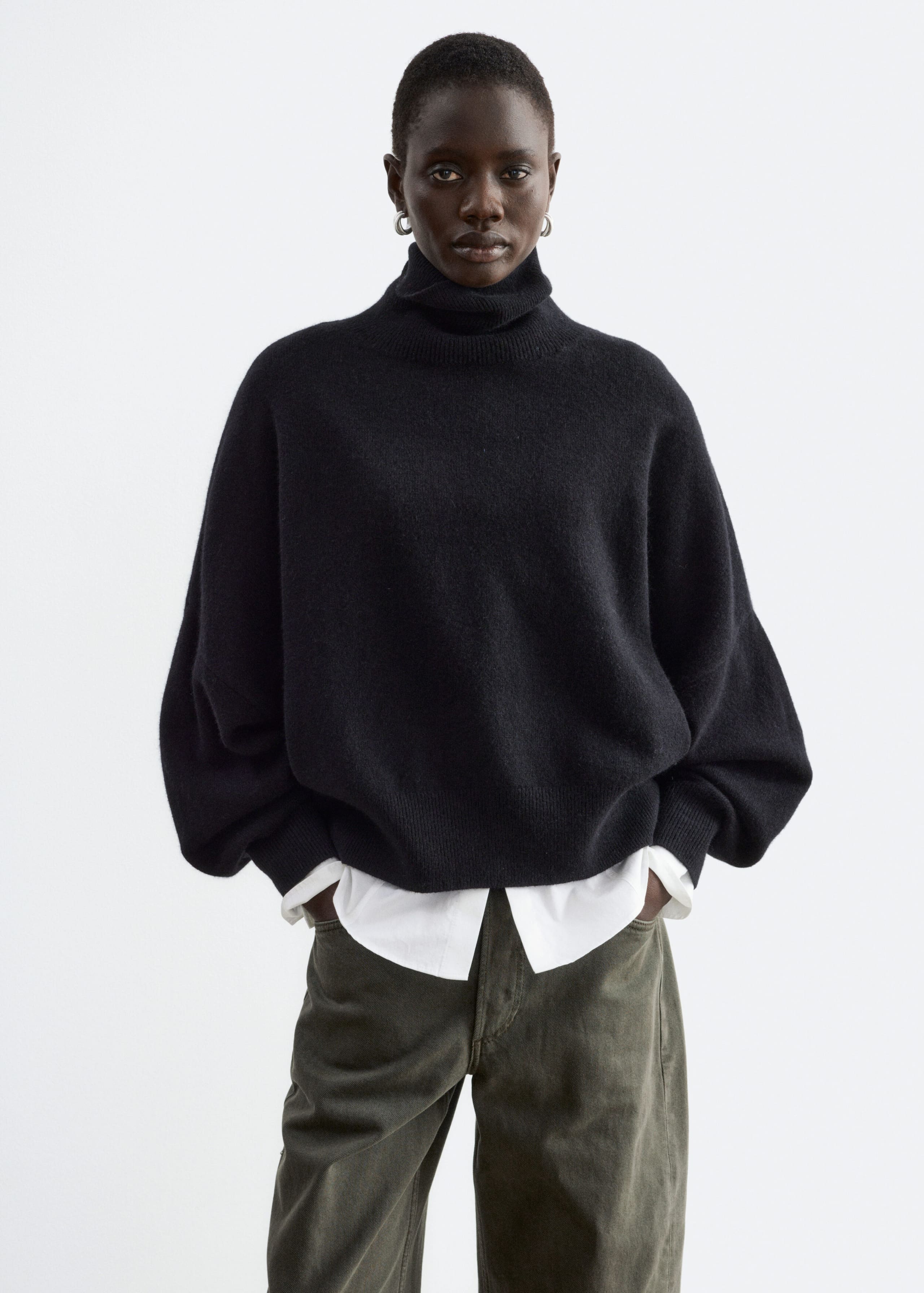 An oversized black jumper featuring a folded rib turtleneck and dropped armholes is being worn by the model. The jumper is layered over a white collared shirt and styled with olive green trousers, with the model resting their hands in the pockets.