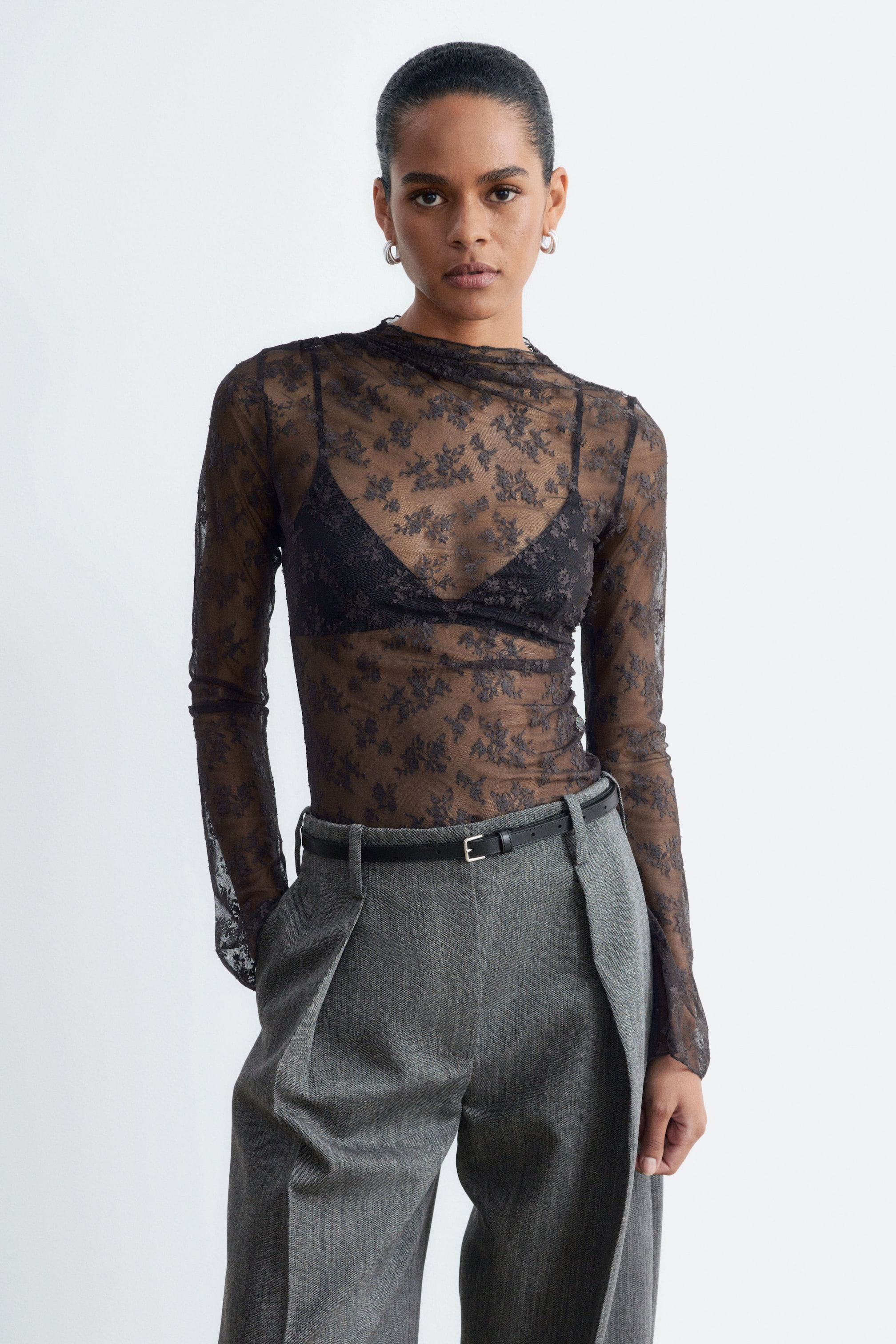 Visualizza immagine più grande: Una top in pizzo floreale nero trasparente, con collo alto aderente e arricciatura asimmetrica sulla spalla, indossata dalla modella. La top a maniche lunghe è sovrapposta a un bralette nero e infilata in pantaloni grigi plissettati.