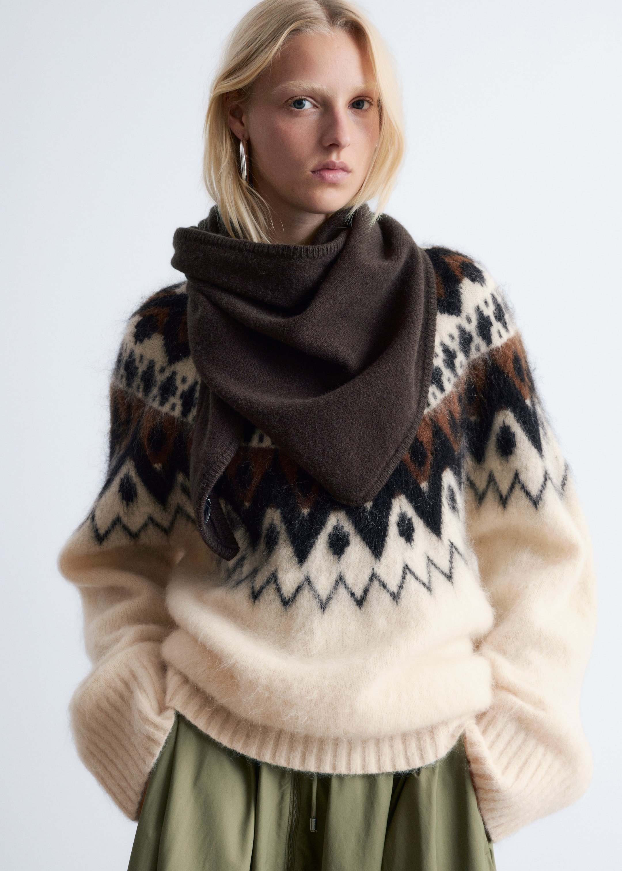 Agrandir l'image: Un pull Fair Isle de couleur crème, avec des motifs géométriques noirs, brun foncé et tan sur une maille duveteuse, est porté en coupe oversize avec des manches longues et un ourlet côtelé, superposant un pantalon vert olive. Une écharpe en maille brun foncé est drapée autour du cou et de la poitrine supérieure, couvrant partiellement le motif du pull.