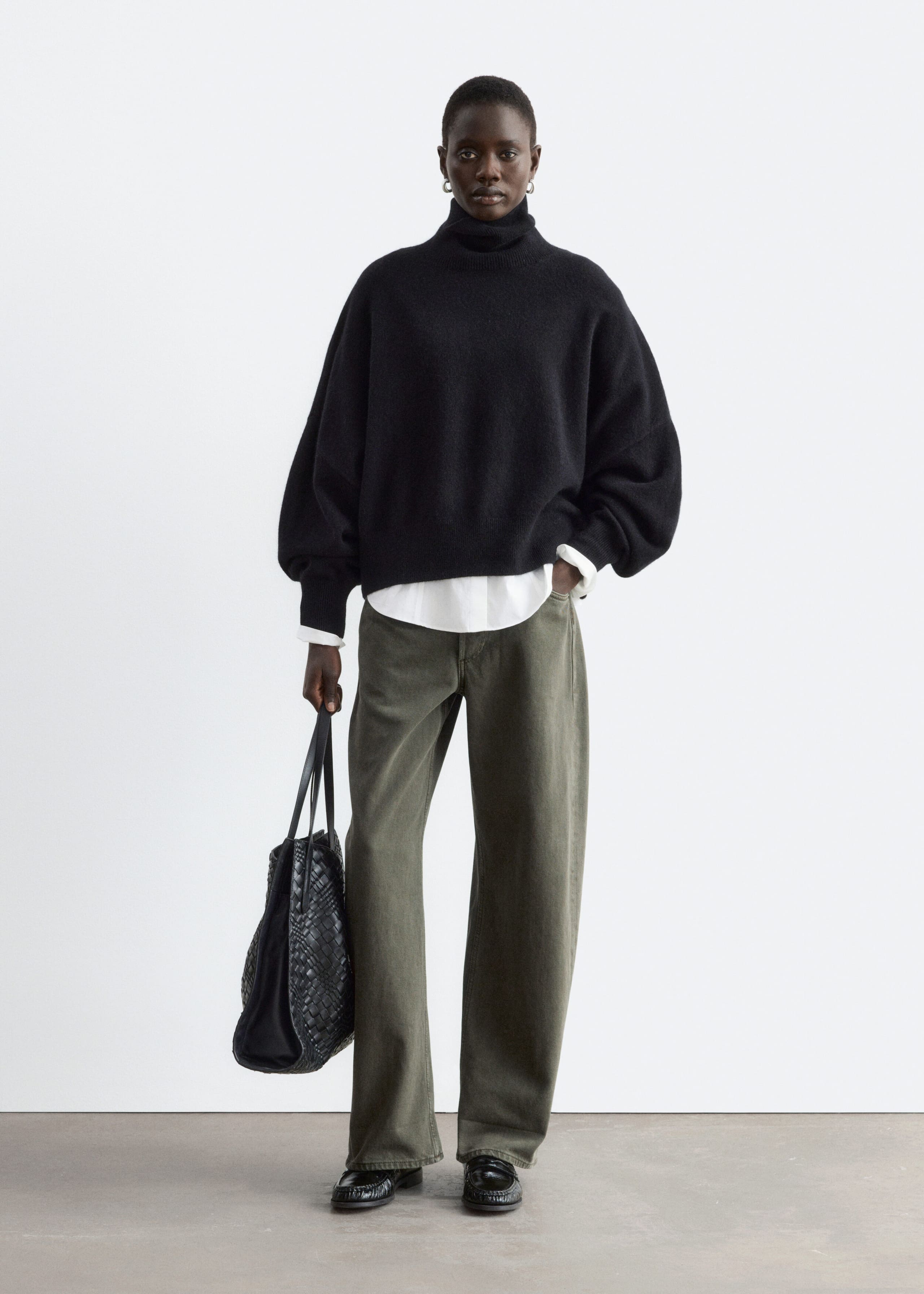 An oversized black knitted jumper featuring a folded rib turtleneck and dropped shoulders is being worn by the model. The jumper is layered over a white collared shirt and styled with olive-green trousers, while a black woven tote bag is being held in one hand.