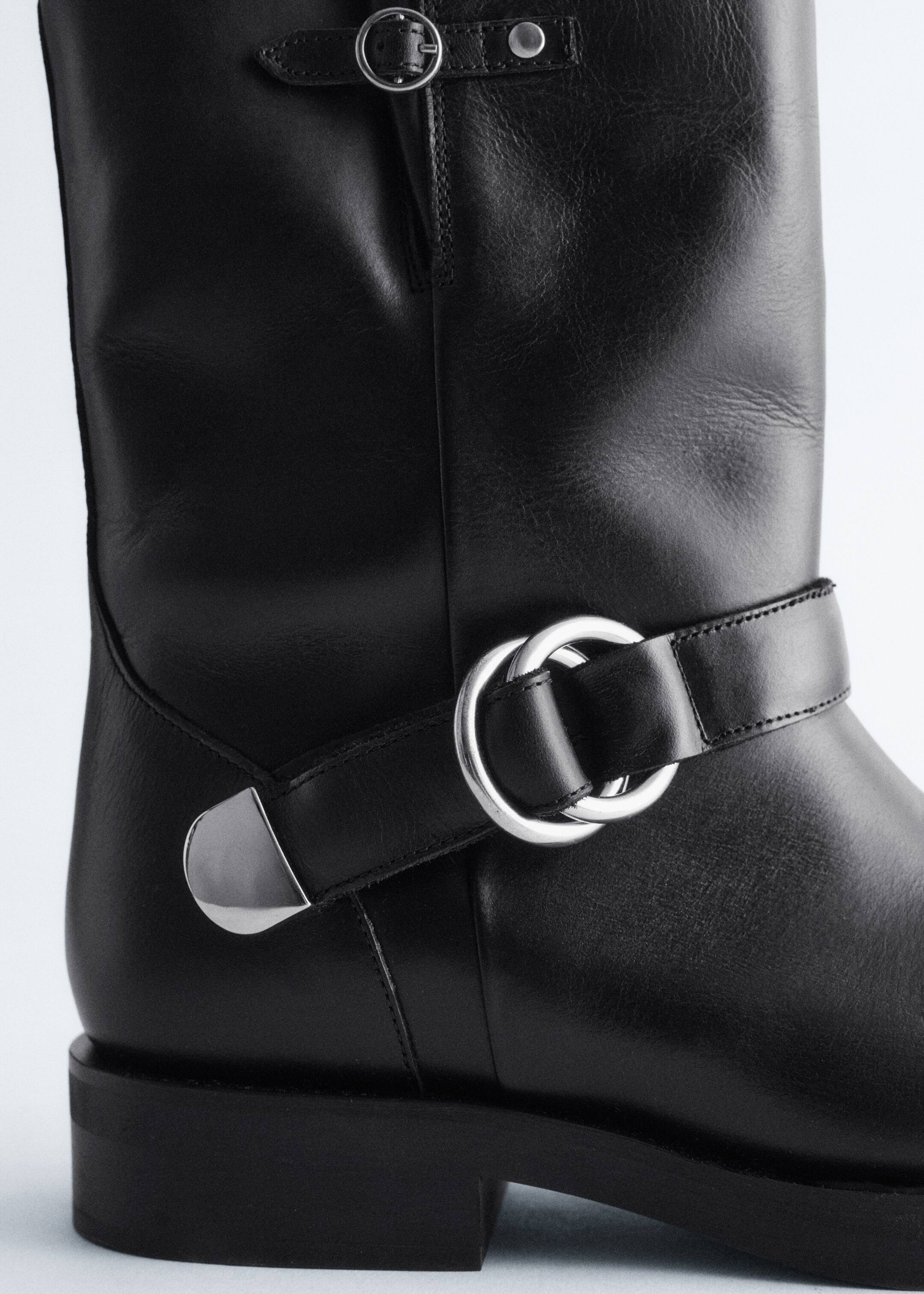 Agrandir l'image: Une botte en cuir noir est présentée en gros plan, vue de côté, mettant en valeur son dessus en cuir noir lisse. Elle est ornée de ferrures en métal argenté, comprenant une petite sangle à boucle en haut et une sangle plus large avec deux anneaux entrelacés et un accent triangulaire entourant la cheville, le tout reposant sur une semelle noire épaisse avec un talon bloc.