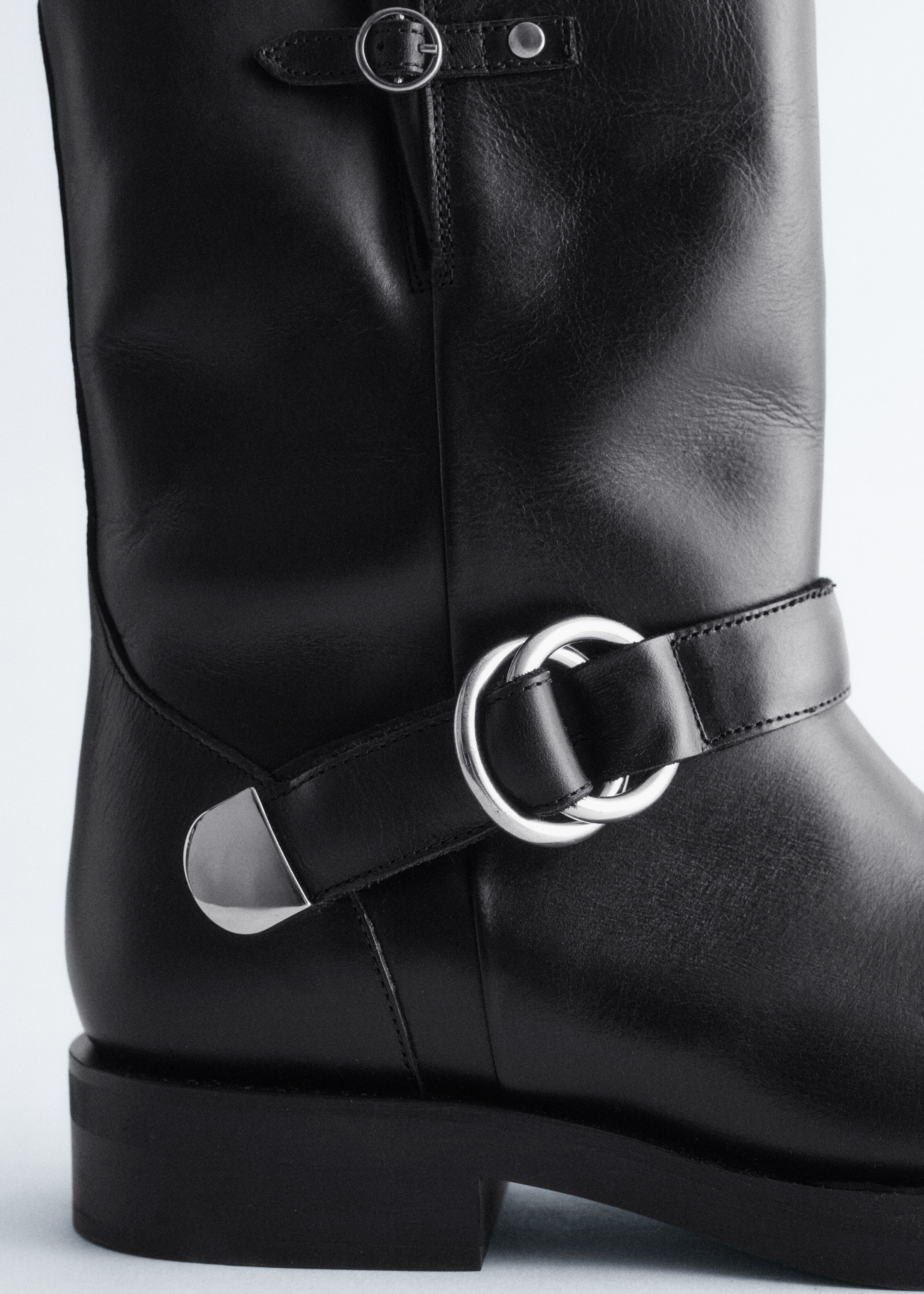 View larger image: A black leather boot is presented in a close-up, side view, showcasing its smooth black leather upper. It is detailed with silver-toned metal hardware, including a small buckle strap at the top and a wider strap with two interlocking rings and a triangular accent encircling the ankle, all resting on a chunky black sole with a block heel.