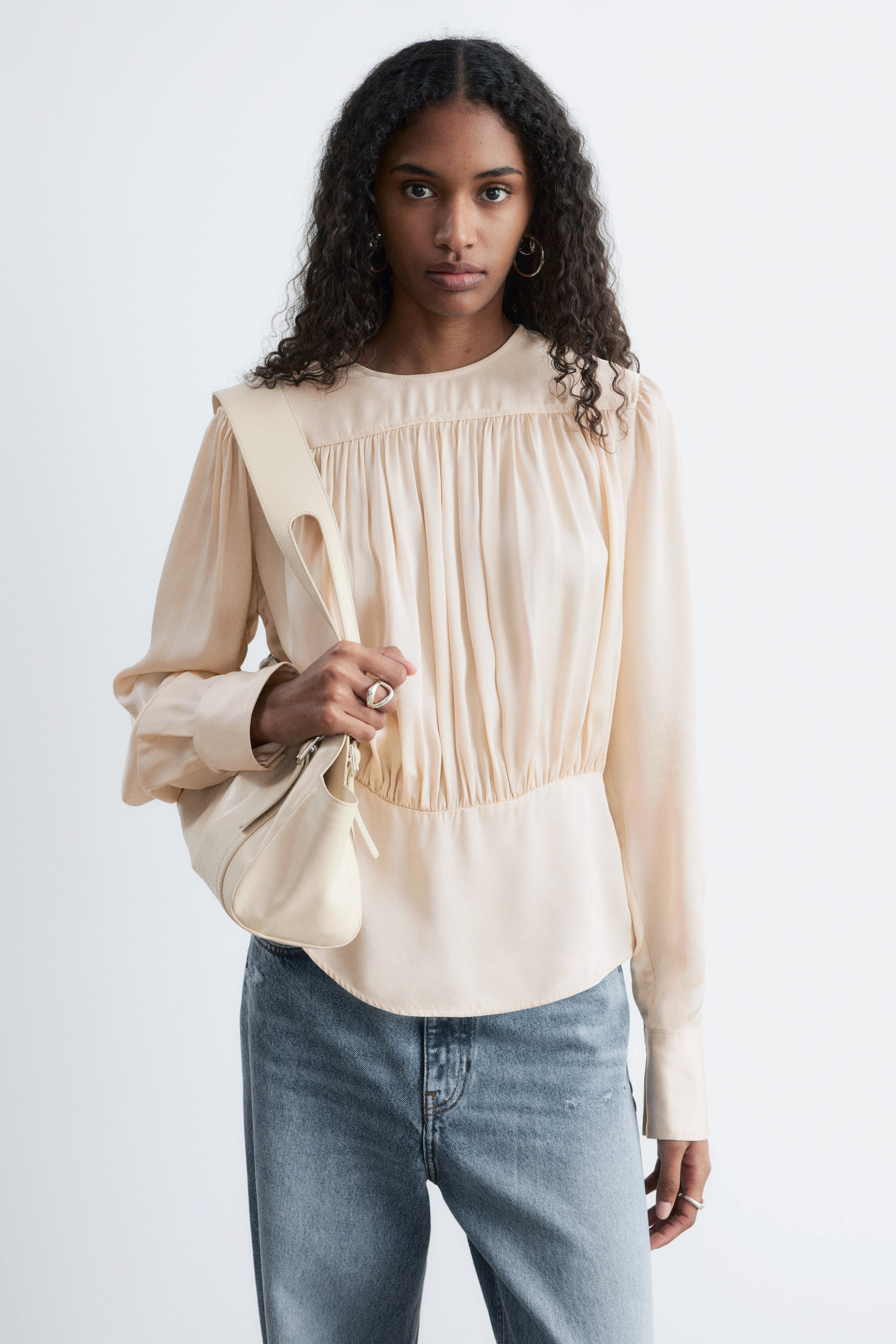 Größeres Bild ansehen: Eine blasse Pfirsichbluse mit Rundhalsausschnitt und langen Ärmeln mit Knopfmanschetten. Sie präsentiert Raffungsdetails über Brust und Taille, die eine figurbetonte Silhouette mit abgerundetem Saum schaffen.